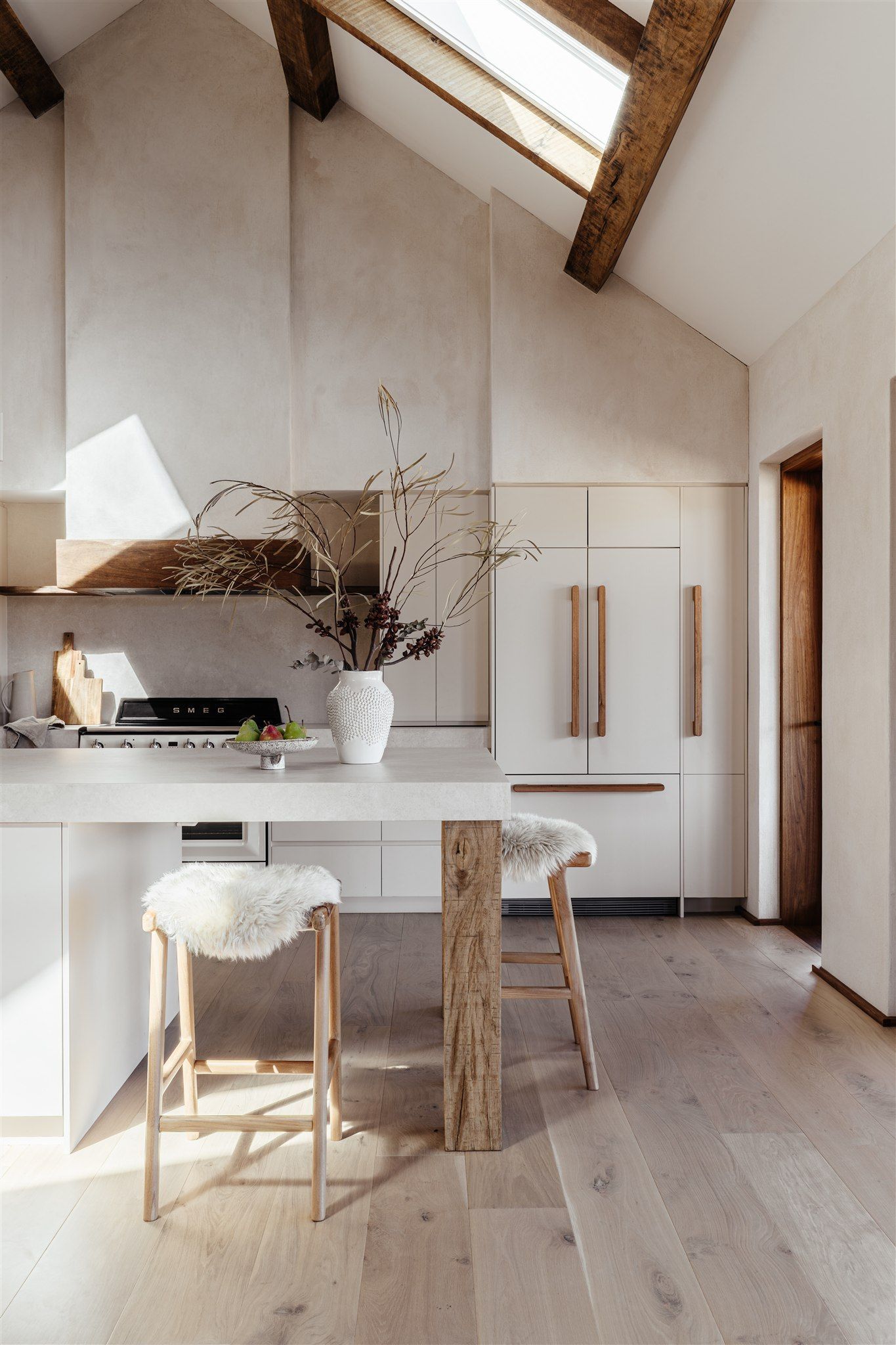 Modern kitchen with white walls, wooden ceiling beams, a central island with bar stools, and warm natural sunlight.