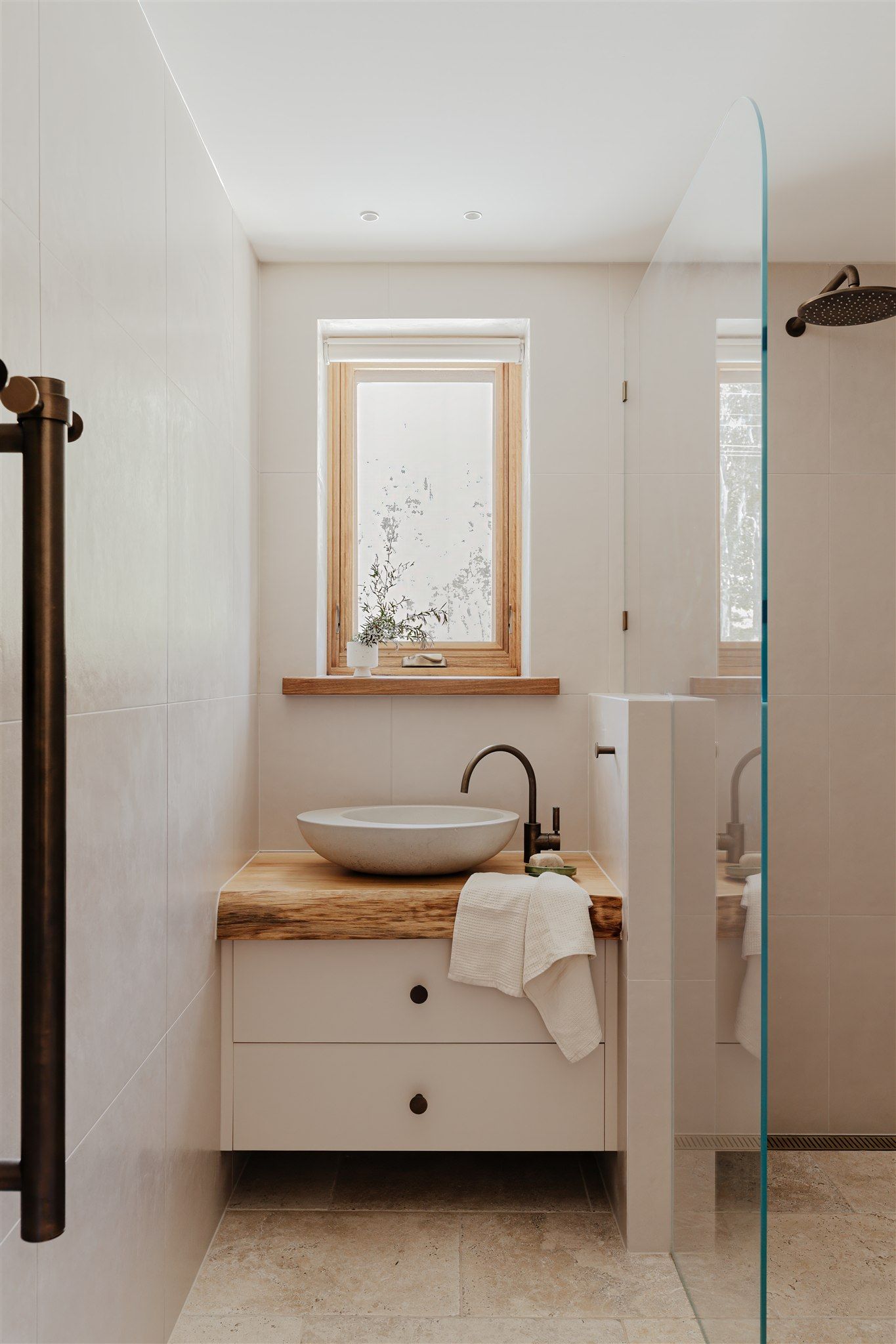 A modern, neutral-toned bathroom featuring a stone vessel sink on a rustic wooden vanity, a window, and a glass shower.