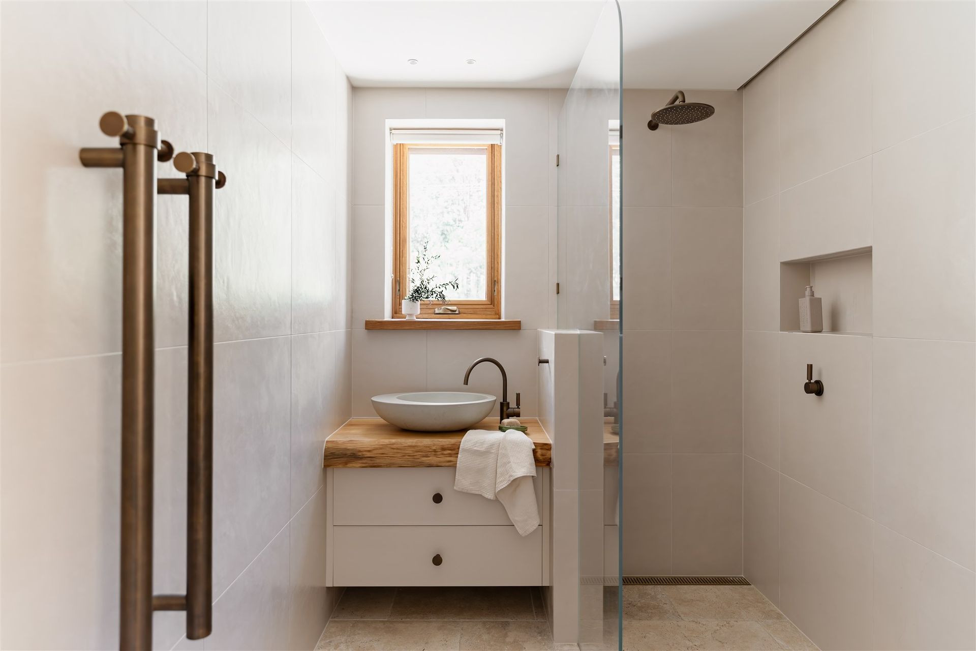 A modern bathroom with a wooden vanity, vessel sink, walk-in glass shower, and bronze towel rails on a white tiled wall.