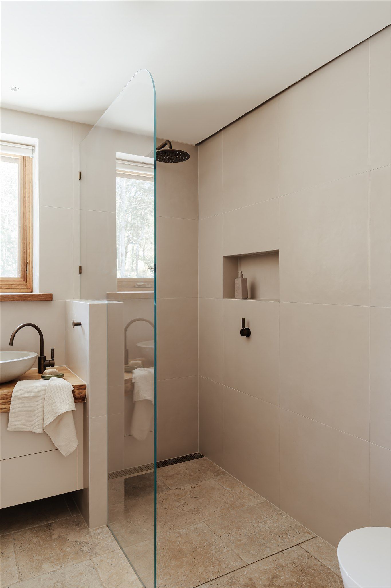 Modern bathroom with a glass-enclosed shower, beige tiled walls, a wooden vanity, and a black faucet.