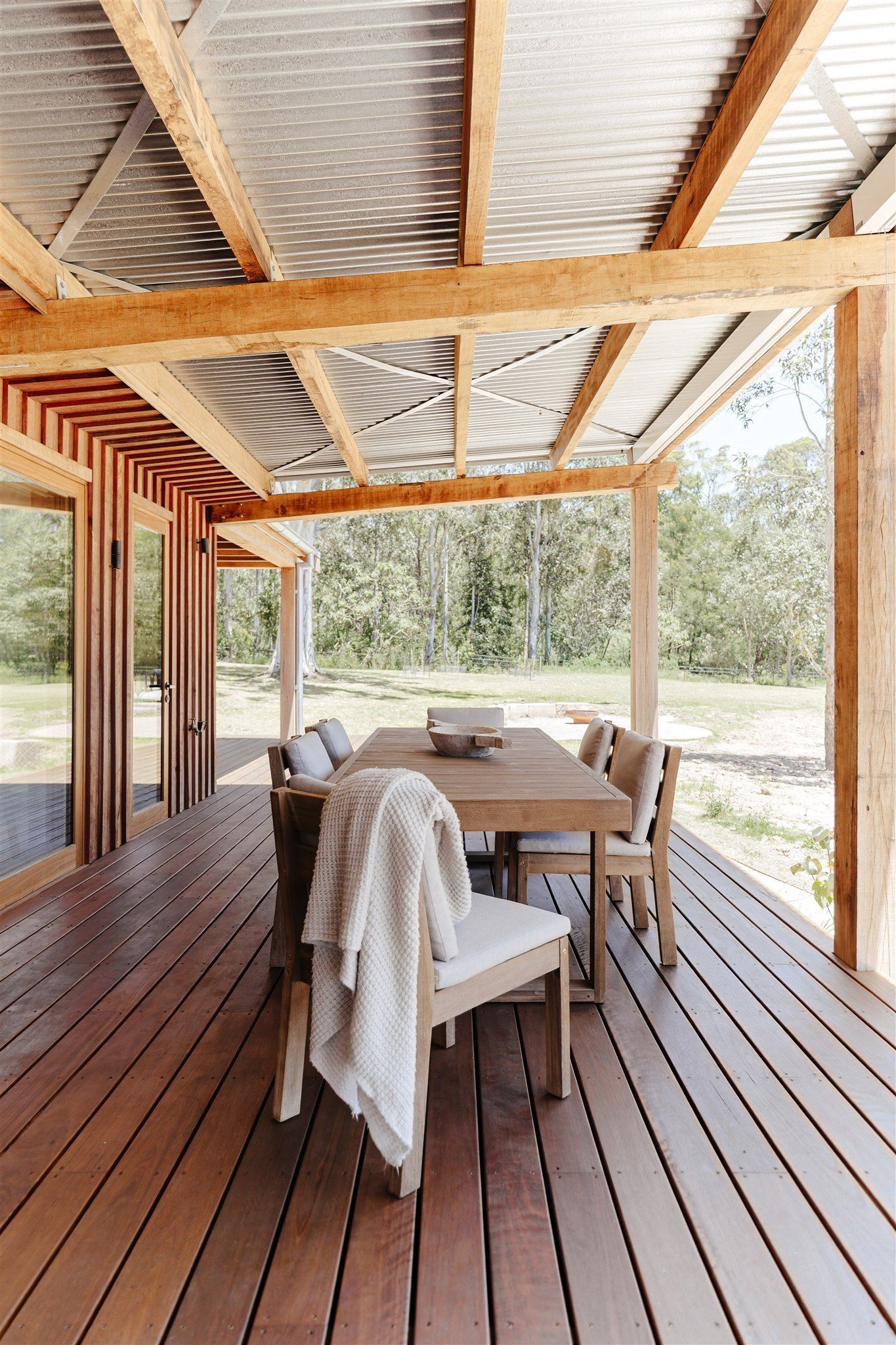Outdoor wooden deck featuring a dining table and chairs with a white throw blanket, under a corrugated metal roof.