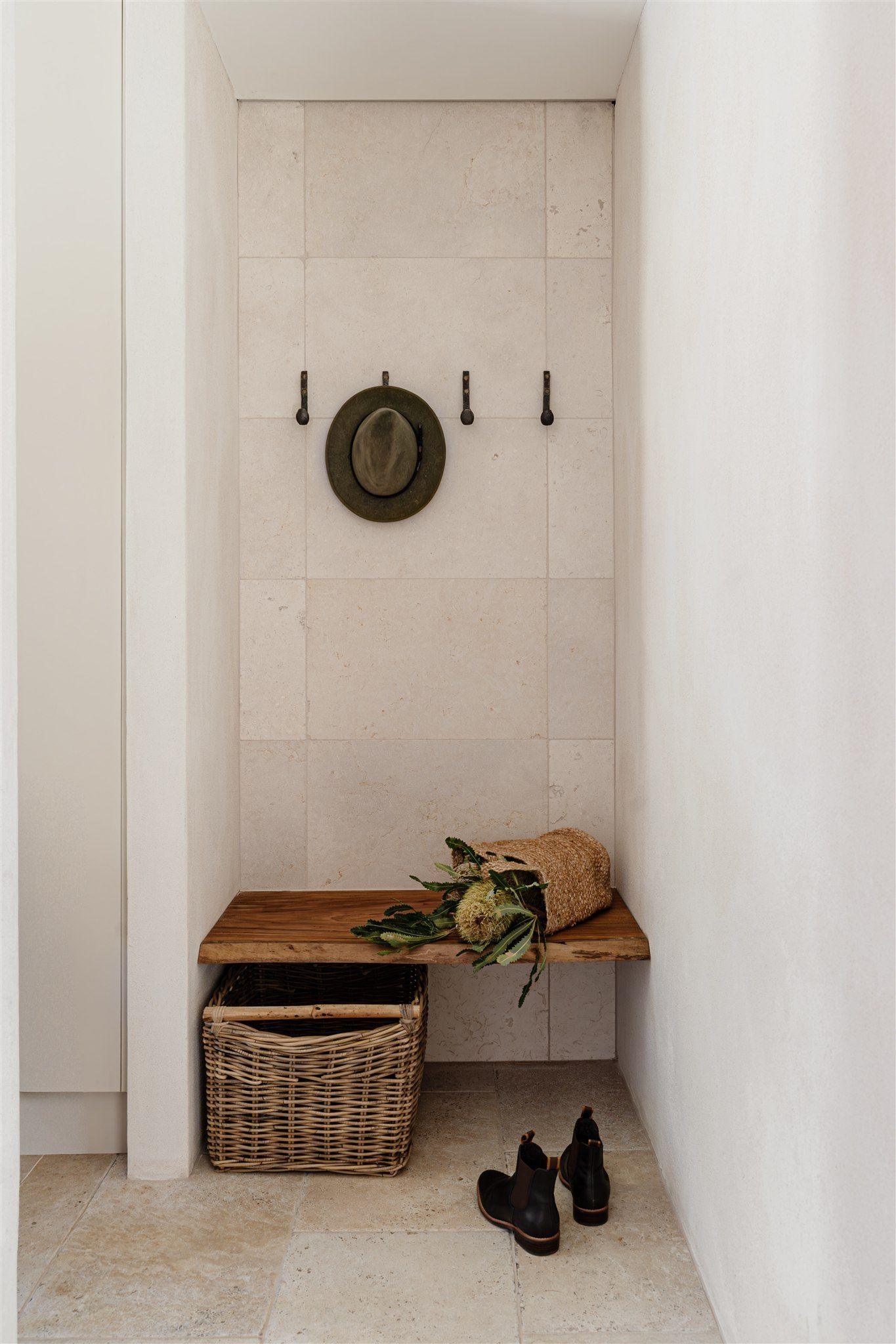 A minimalist entryway nook with a stone-tiled wall, four coat hooks, a wooden bench, a woven basket, and a pair of boots.