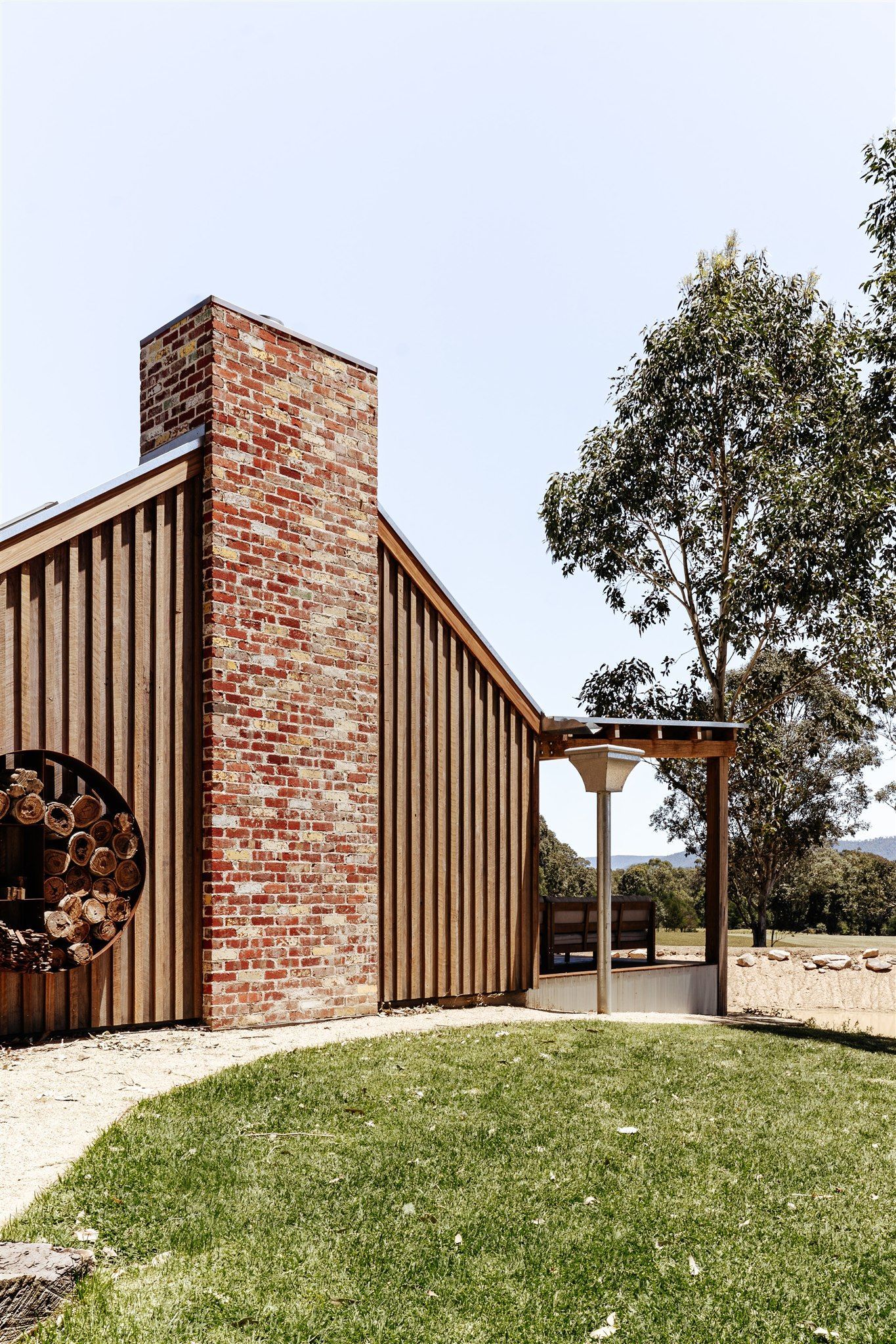 A house with a brick chimney and vertical wood siding sits beside a grass lawn and trees under a clear blue sky.