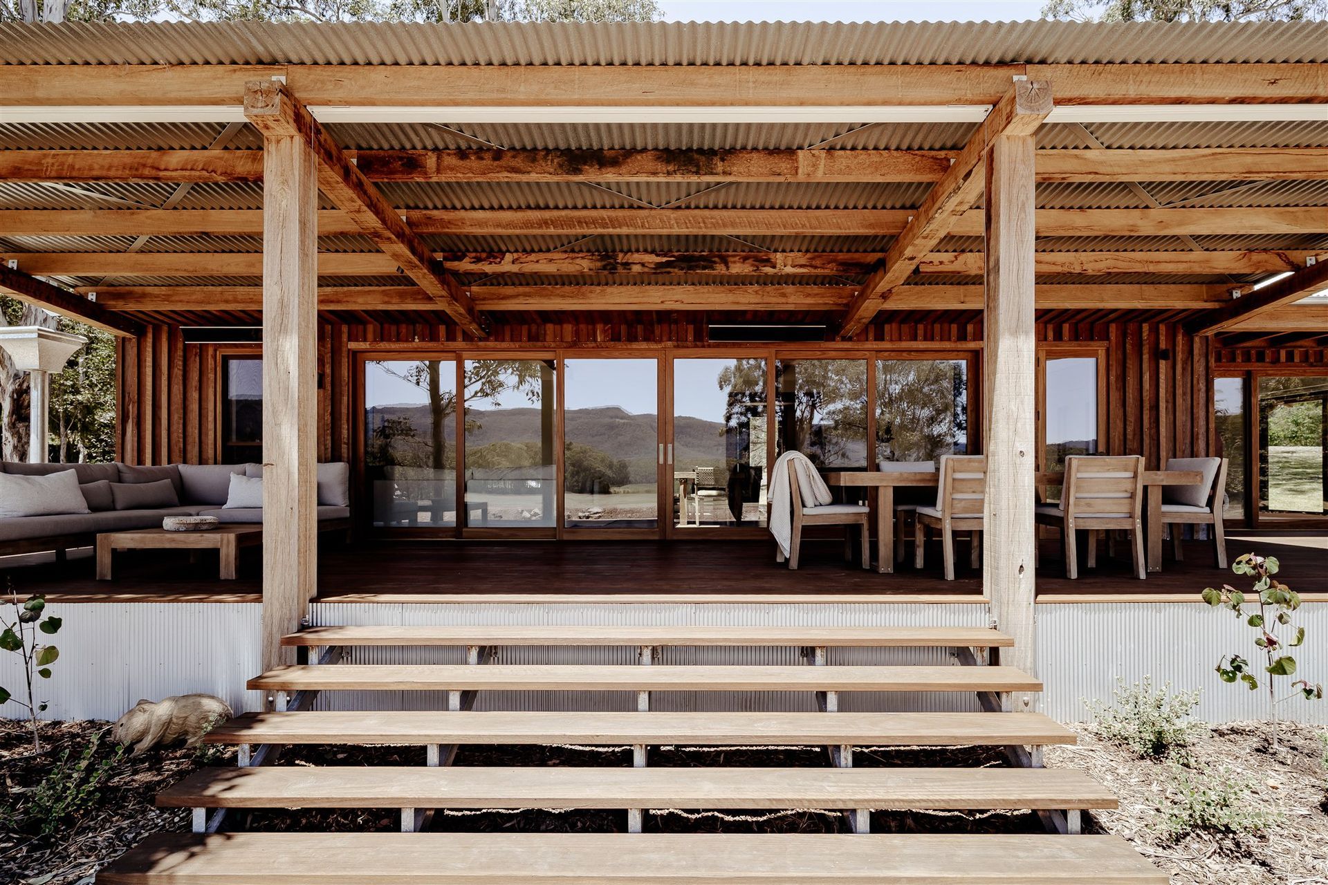 A rustic wooden porch with wide steps, an outdoor sofa, and dining set beneath a corrugated metal roof, facing a landscape.