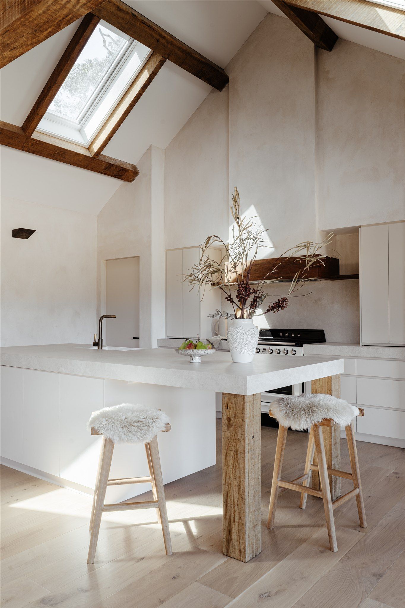 A bright, minimalist kitchen with a large white island, wooden bar stools, exposed ceiling beams, and a skylight.