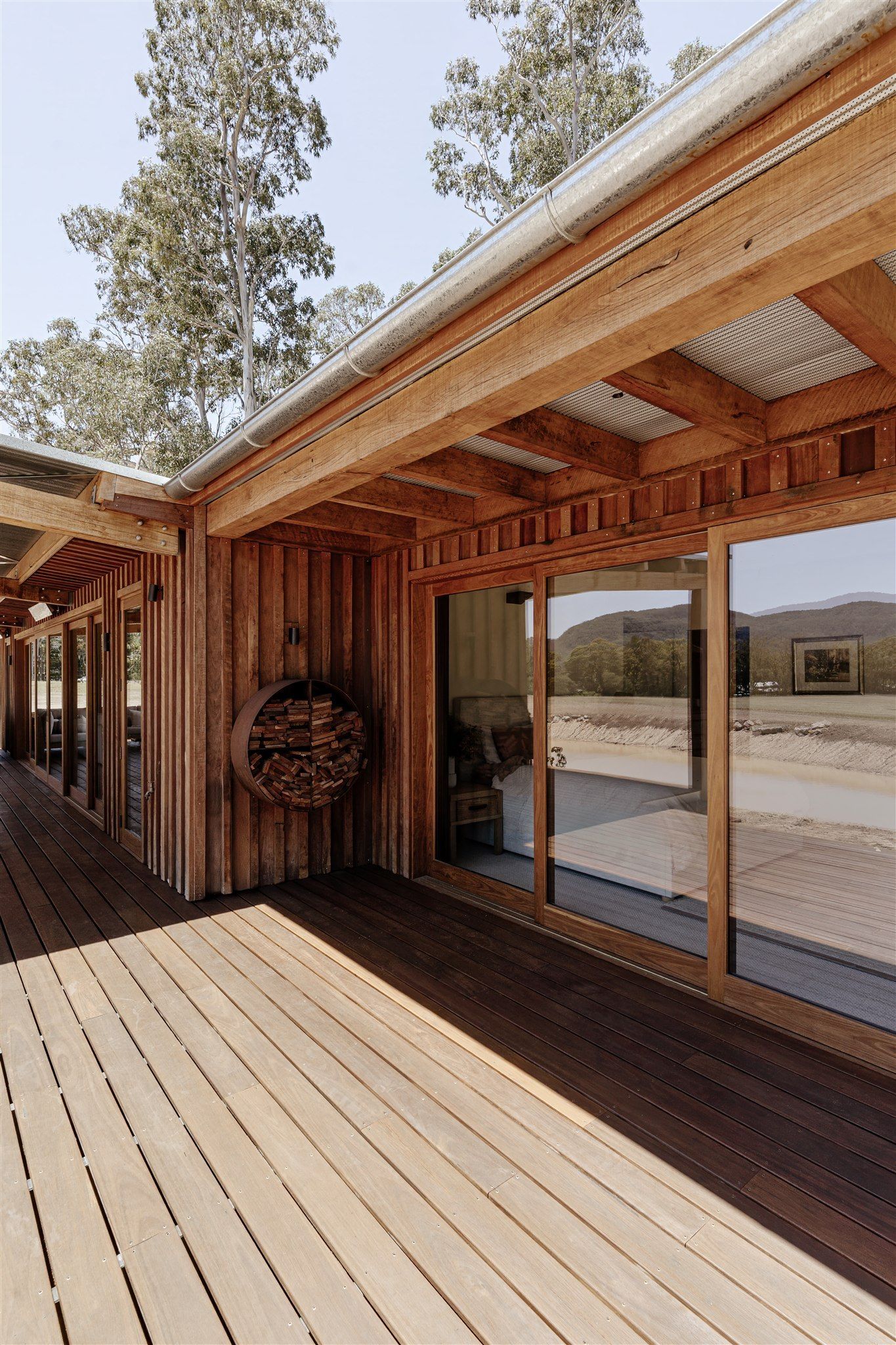 A wooden patio with a mounted log rack, large sliding glass doors, and a rustic beam roof against a bright outdoor scene.