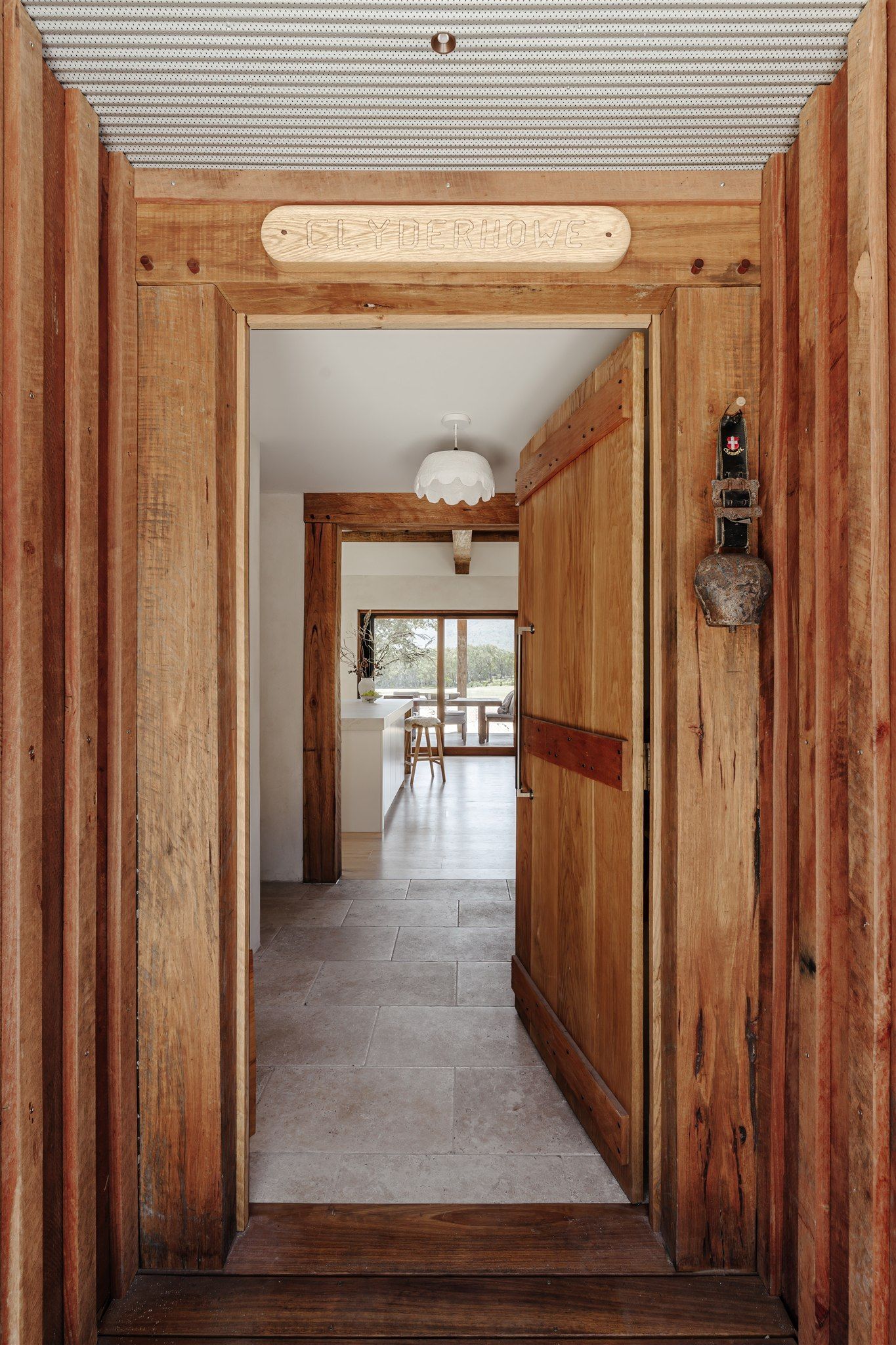An open wooden door framed by rustic timber, looking through a stone-floored hallway to a brightly lit room with a view.