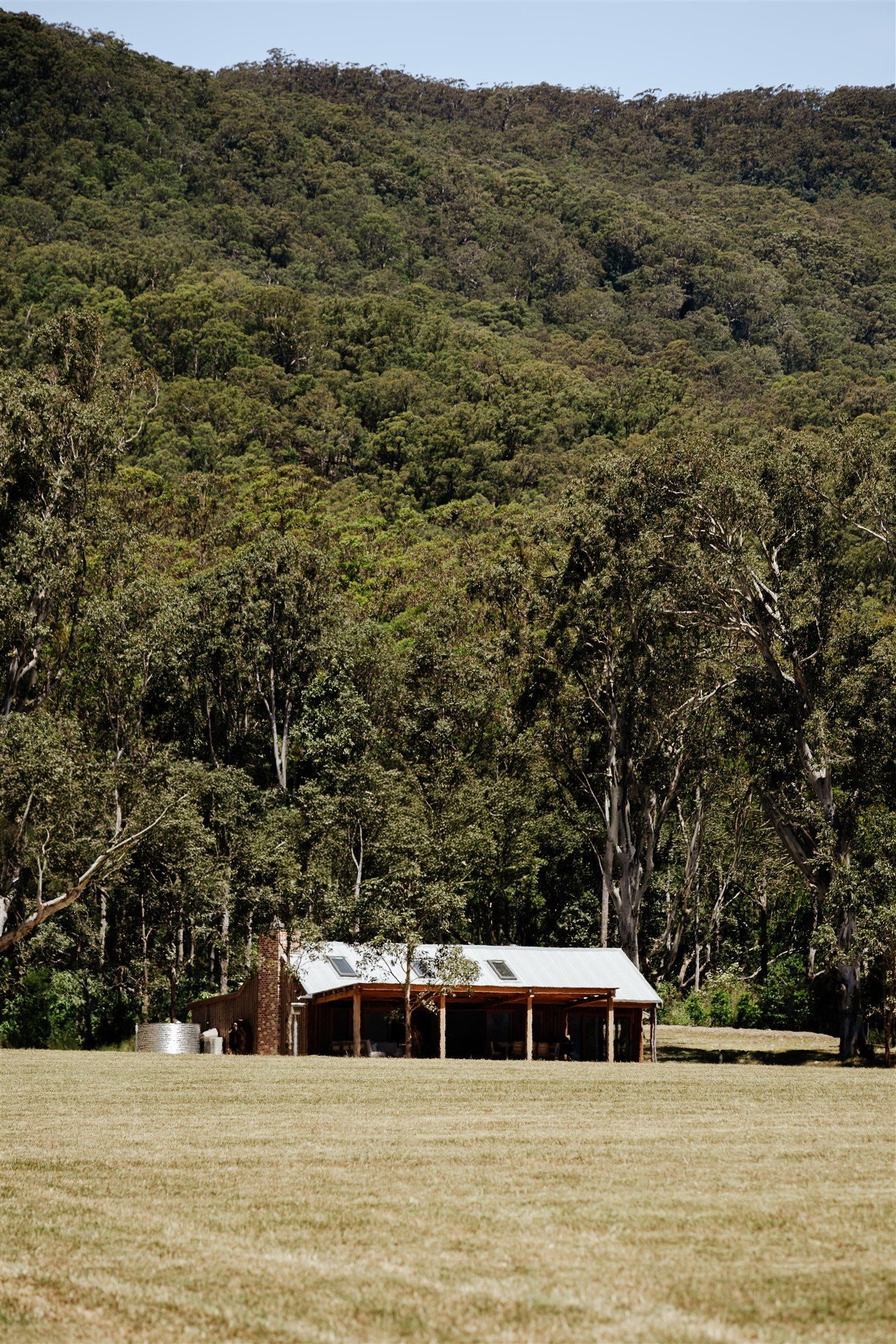 A small, rustic house with a light-colored roof sits in a grassy field backed by a dense, green forested hillside.