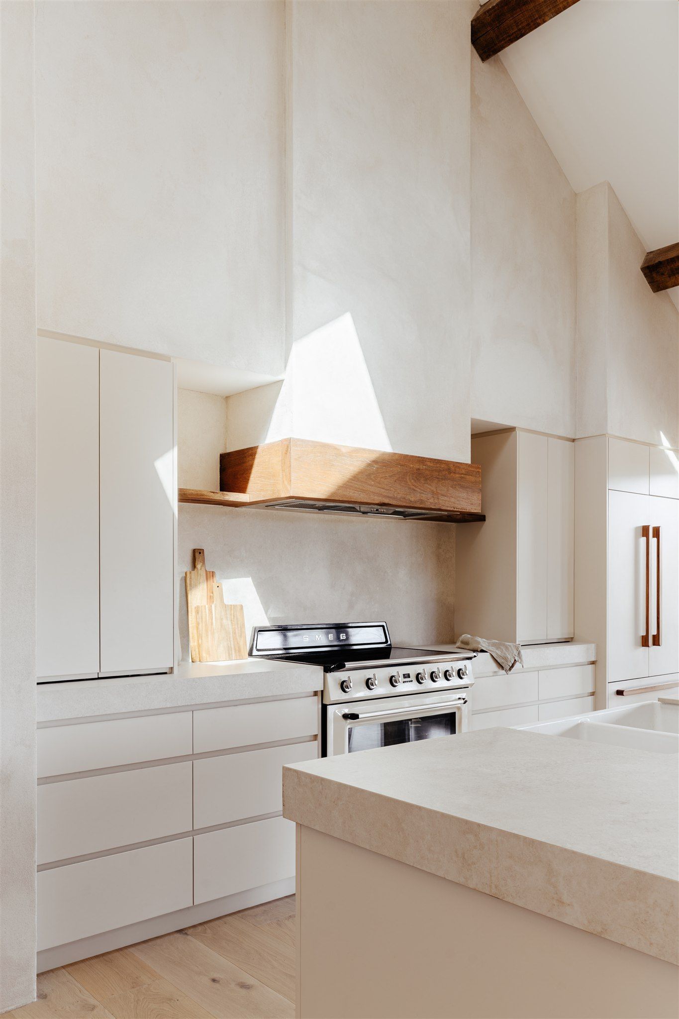A bright, modern kitchen features light-colored cabinets, a minimalist stone range hood with wood trim, and a marble island.