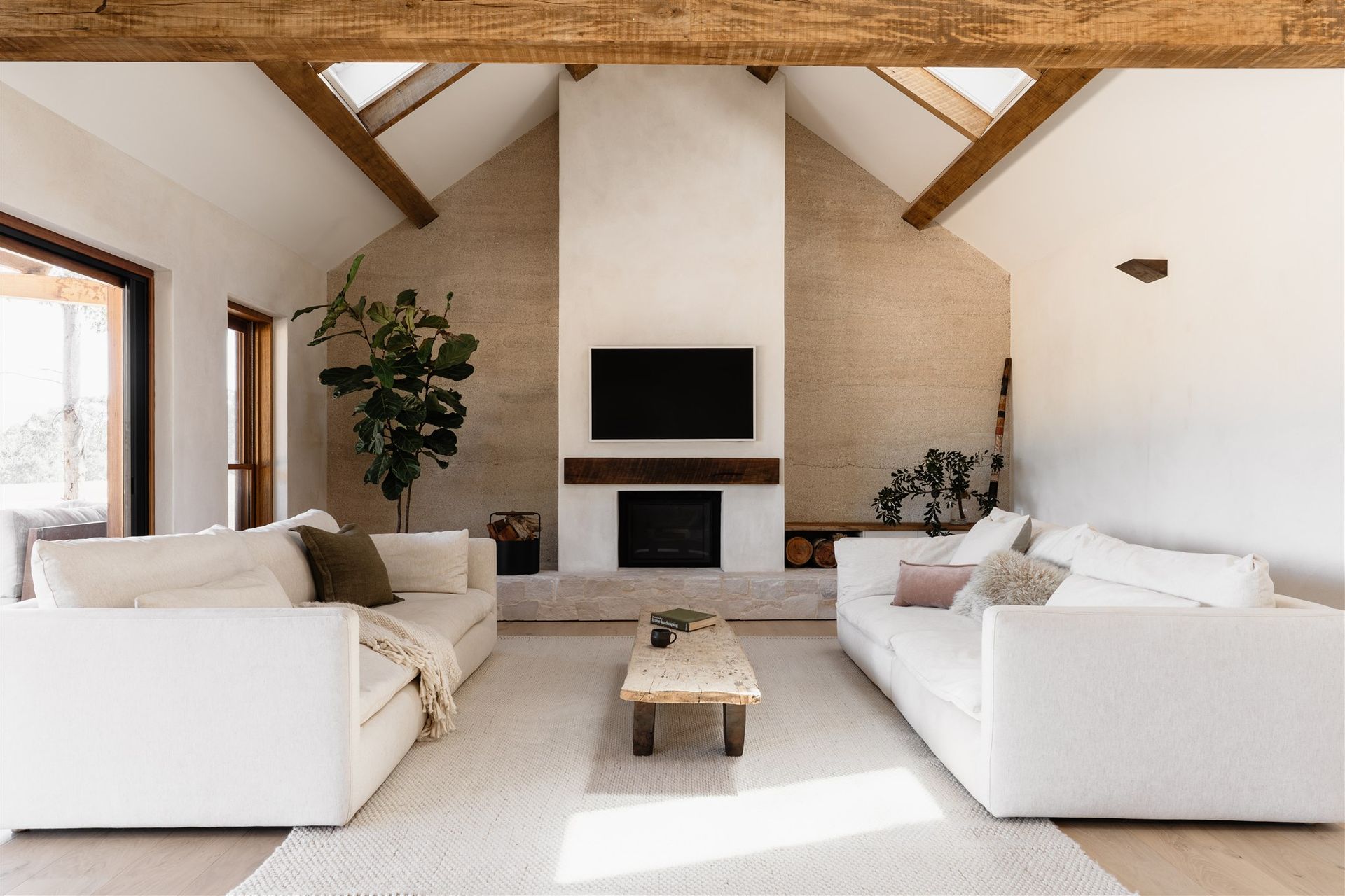 A bright, modern living room with two white sofas, a central fireplace, wooden beams, and textured stone walls.