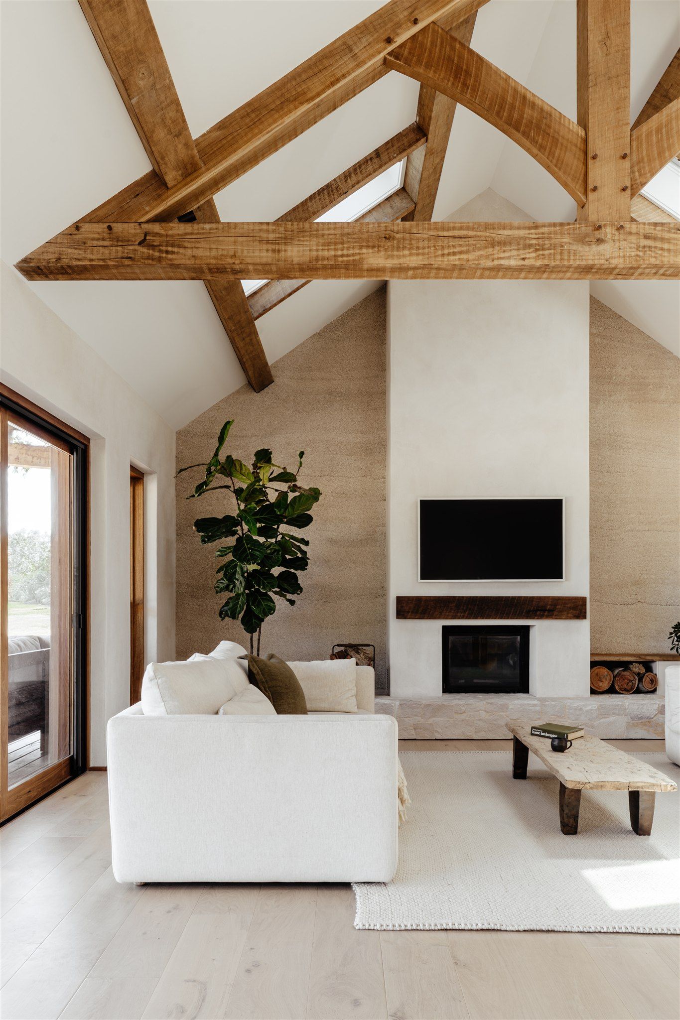 A bright living room with vaulted ceilings, exposed wooden beams, a white sofa, and a stone fireplace with a mounted TV.