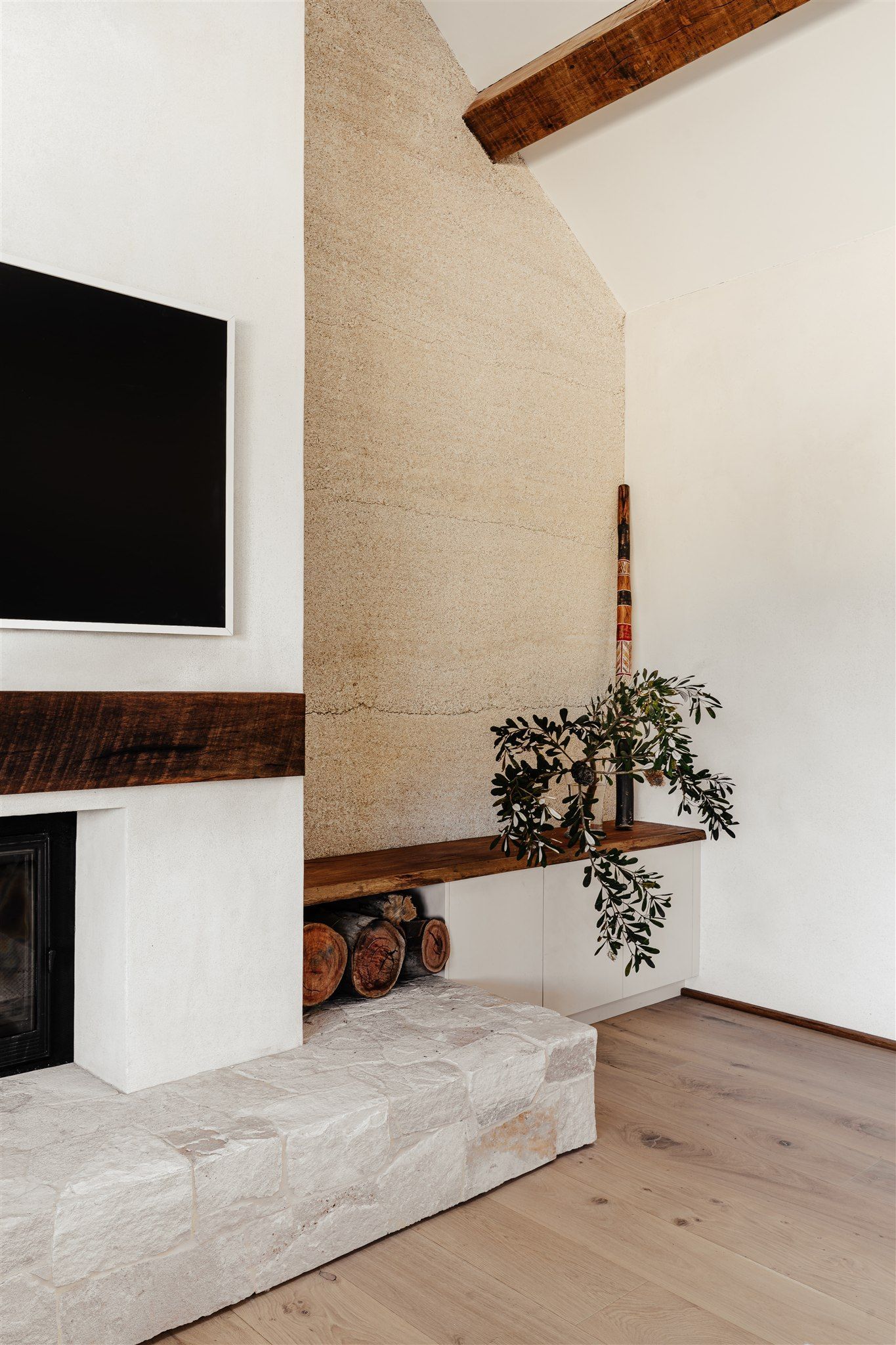 A fireplace with a textured stone wall, wooden mantel, and built-in shelving holding firewood and a small plant.