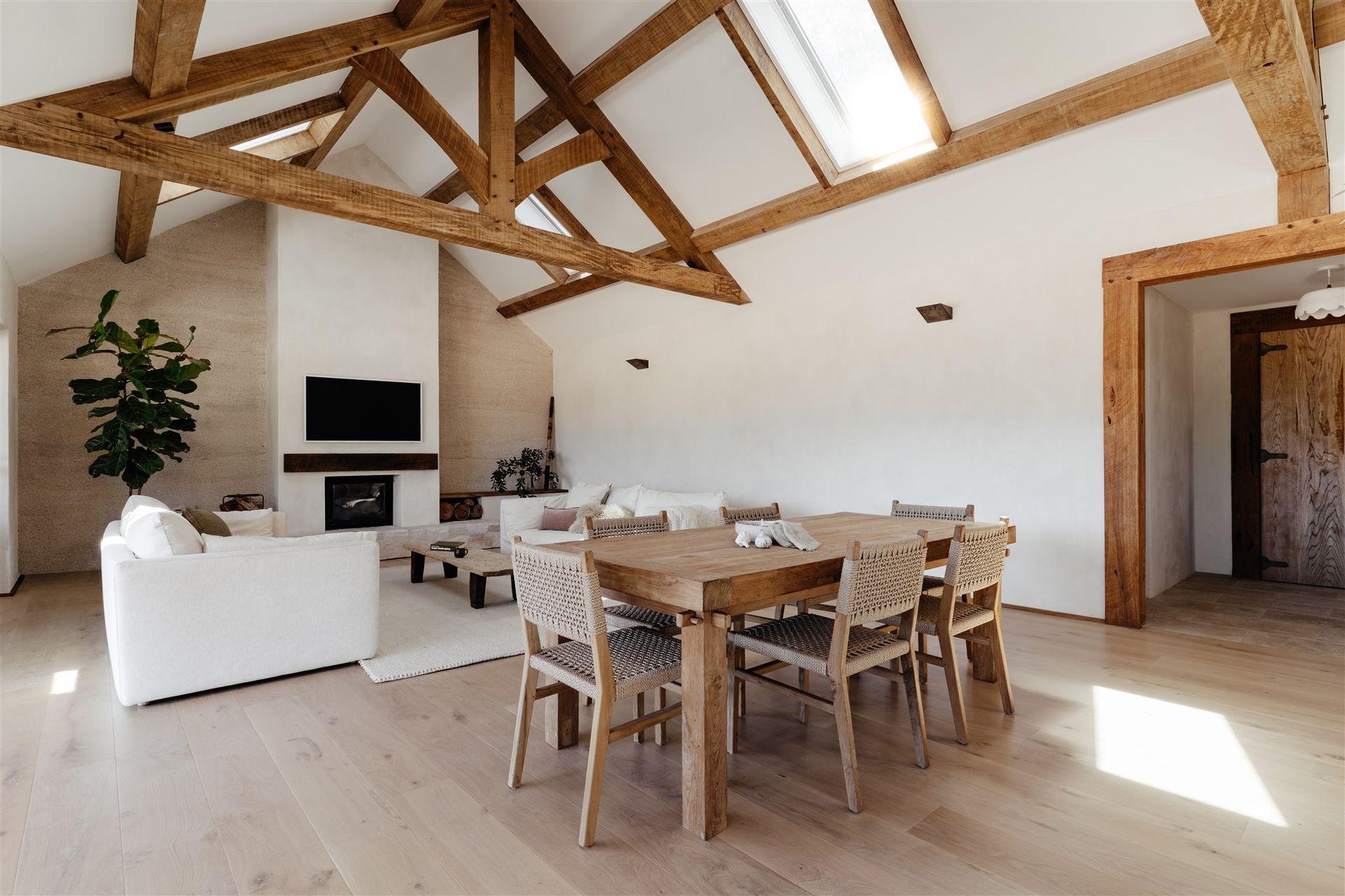 A bright, open-plan living room with a vaulted ceiling, exposed wooden beams, a fireplace, dining area, and white furniture.