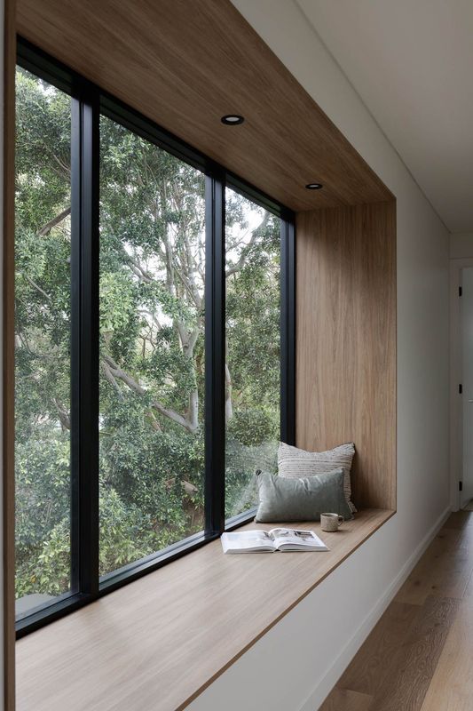 Window seat nook with light wood and black-framed window, view of greenery, cushion, and book.