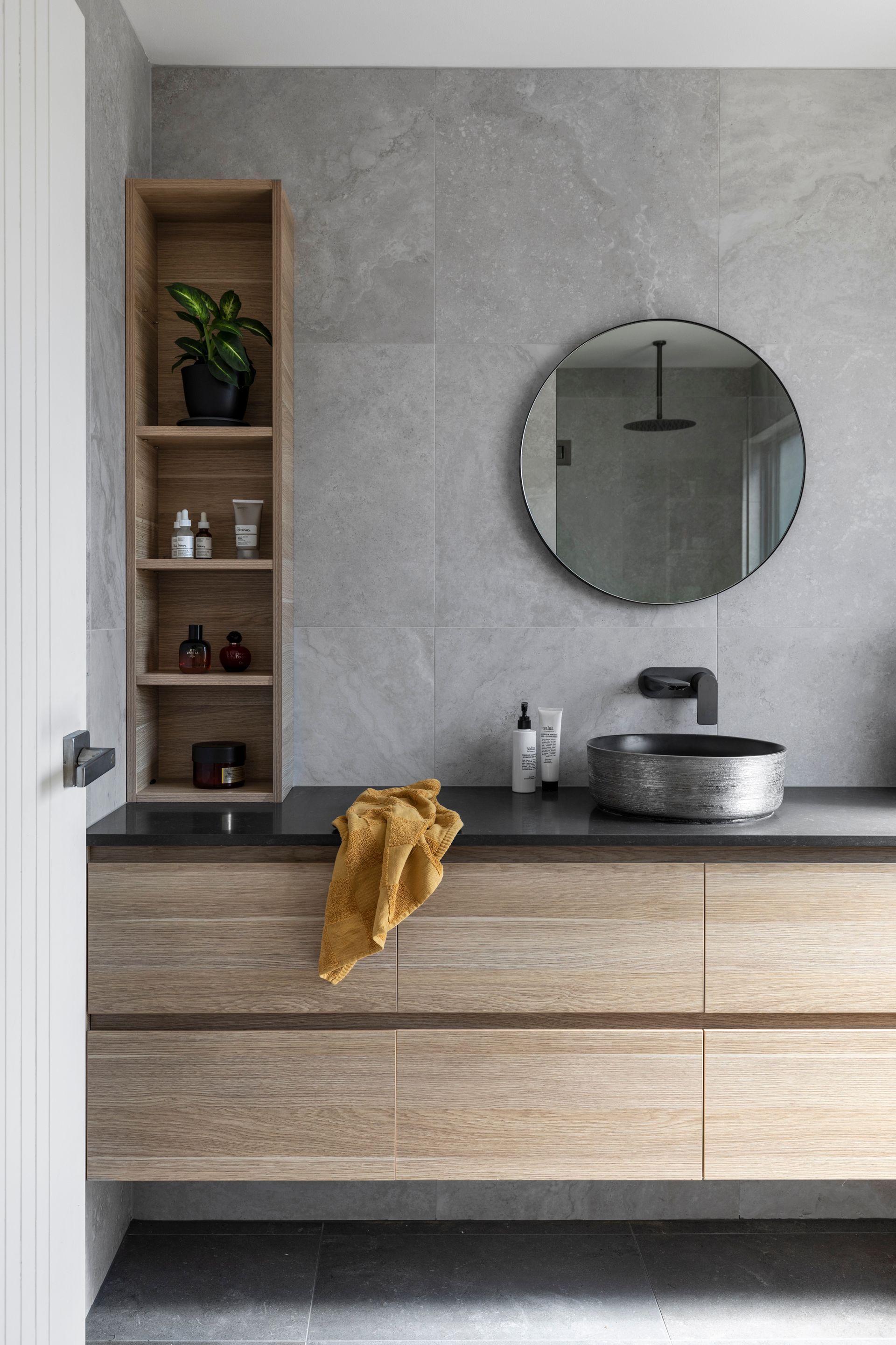 Modern bathroom with wood vanity, gray tile, round mirror, and a woven towel.