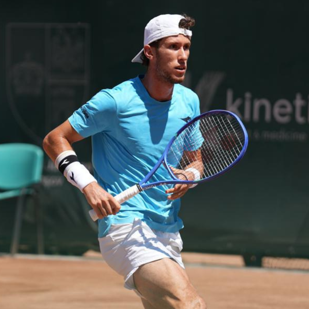 Giocatore di tennis in campo, con berretto bianco, maglietta blu e pantaloncini bianchi, e una racchetta in mano.