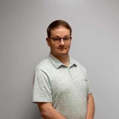 A man wearing glasses and a polo shirt is standing in front of a white wall.