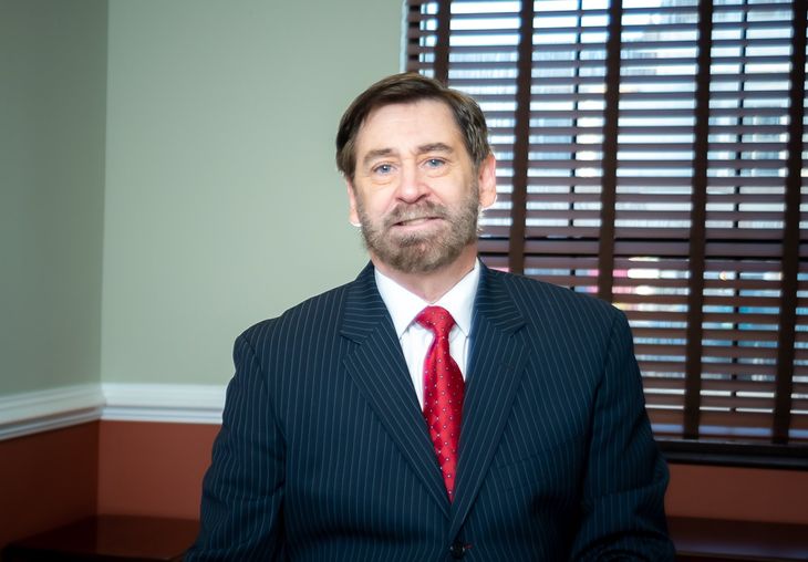 A man in a suit and tie is standing in front of a window with blinds.