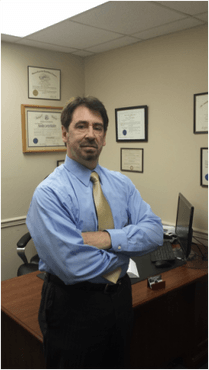 A man in a blue shirt and tie stands in front of a desk with his arms crossed
