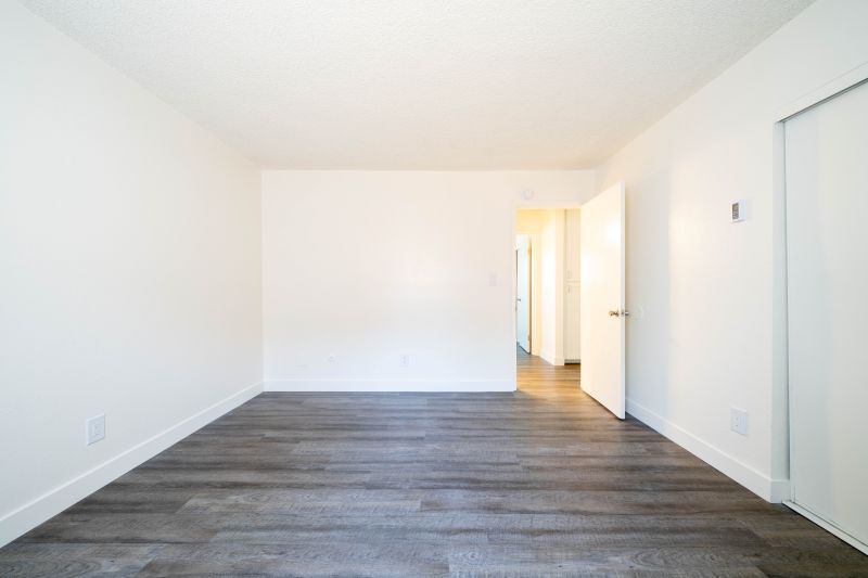 Empty white room with dark wood floors and a bright hallway doorway at the back