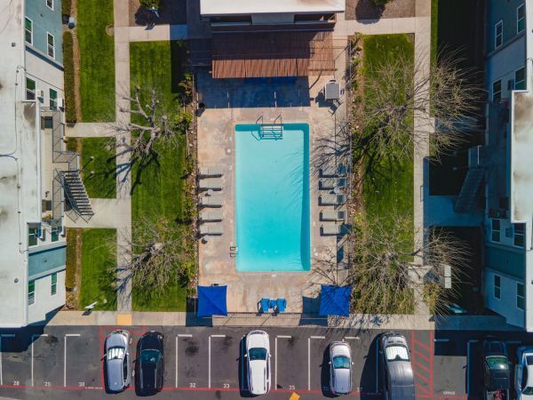 Aerial view of a rectangular outdoor pool between apartment buildings, with lounge chairs and parked cars nearby.