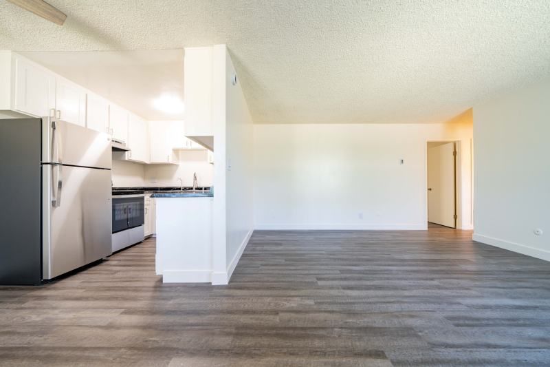 Empty open-plan apartment with kitchen, stainless steel appliances, white walls, and wood-look floors
