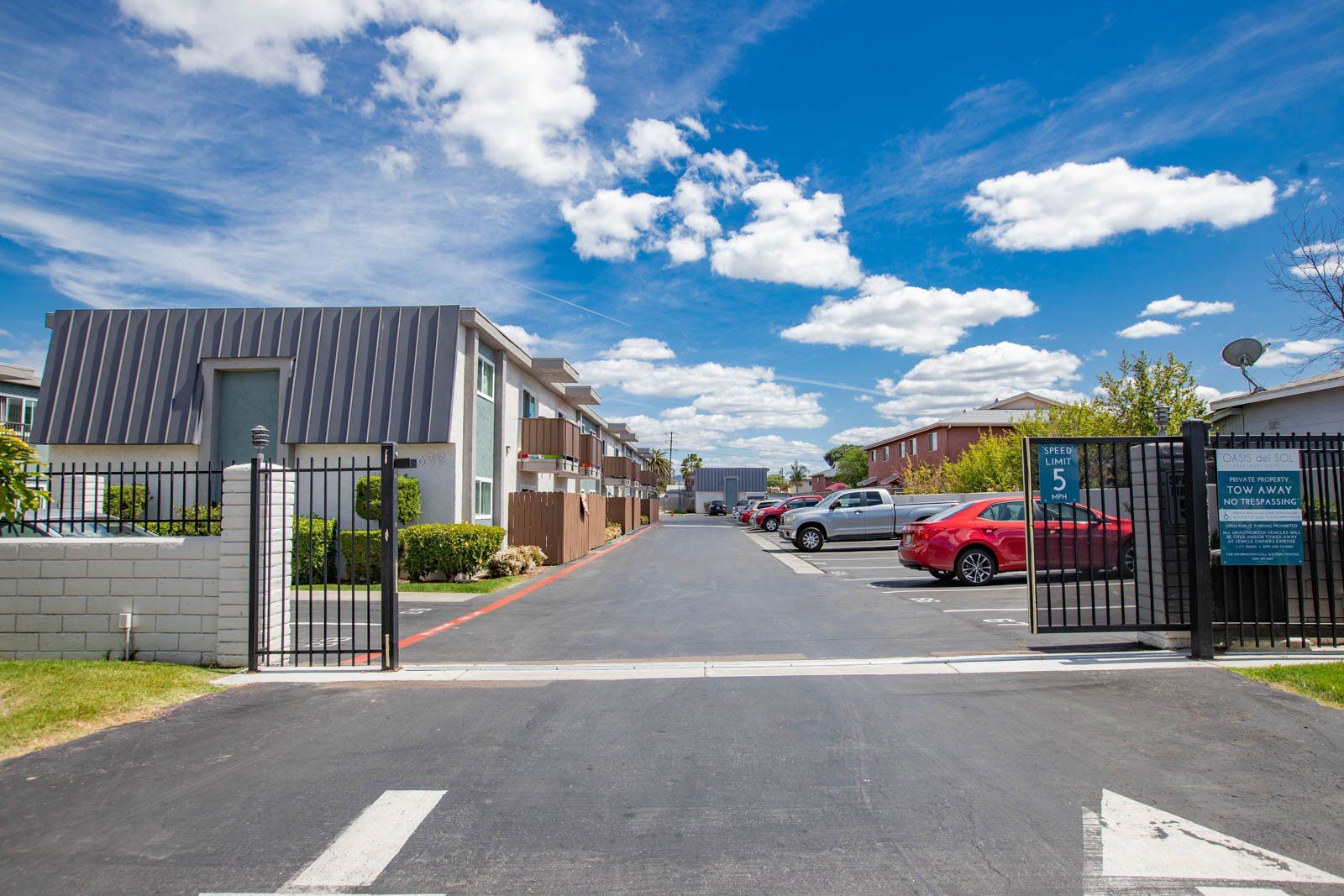 Gated apartment complex entrance with parked cars and a paved driveway under a blue sky