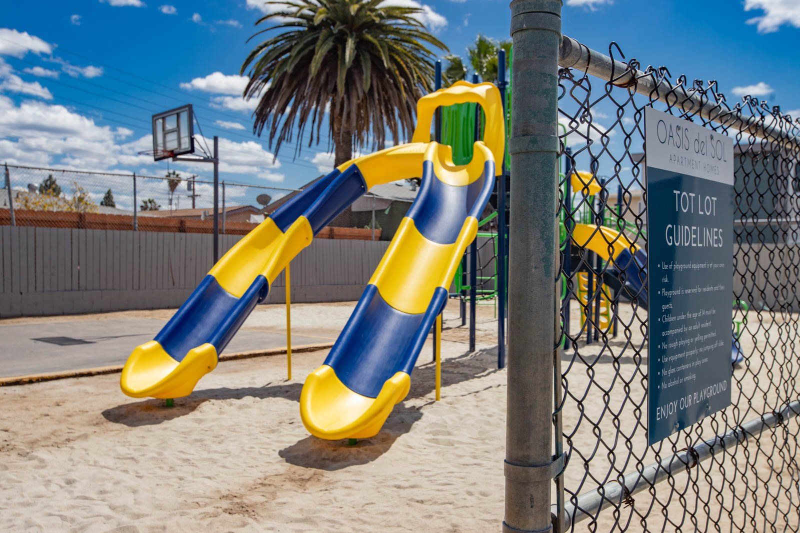 Blue-and-yellow playground slide beside a chain-link fence, with palm trees and a clear sky.