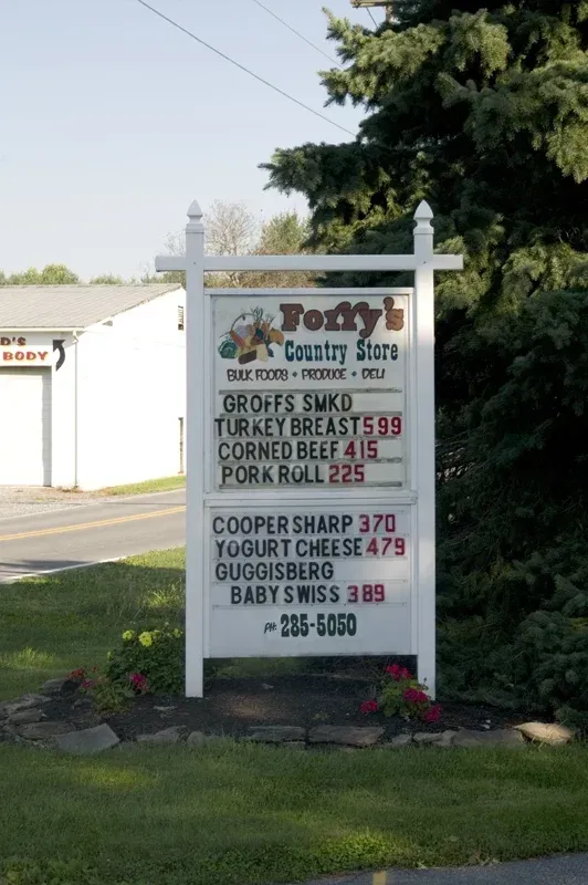 A sign for Foffy's Country Store lists prices for deli meats and cheeses, positioned outdoors near a road and evergreen.