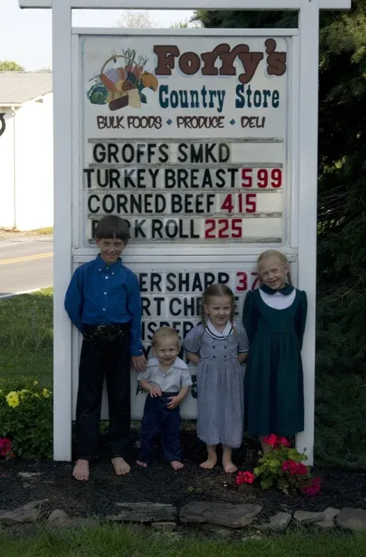 Four children stand barefoot in front of a sign for Foffy’s Country Store, which displays prices for deli meats and cheese.