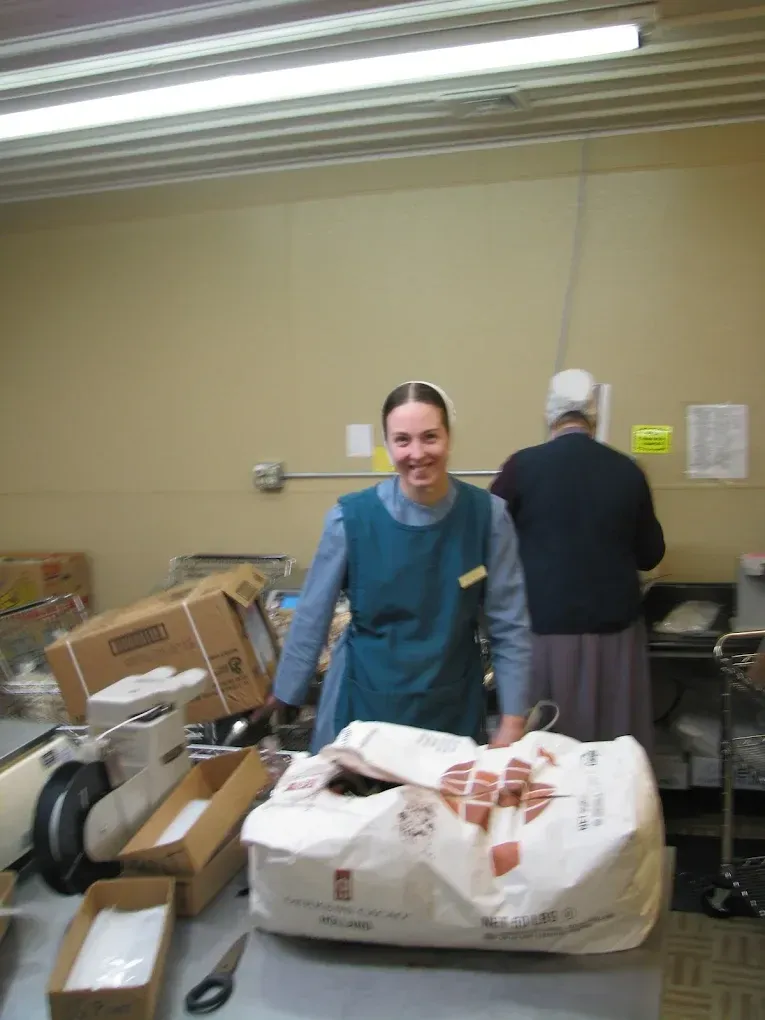 A smiling person wearing a blue apron and head covering stands at a workspace with boxes and a bag of supplies.