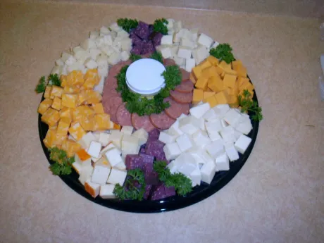 A round party platter featuring segments of cubed yellow and white cheeses, purple cheese, and sliced salami around a dip.