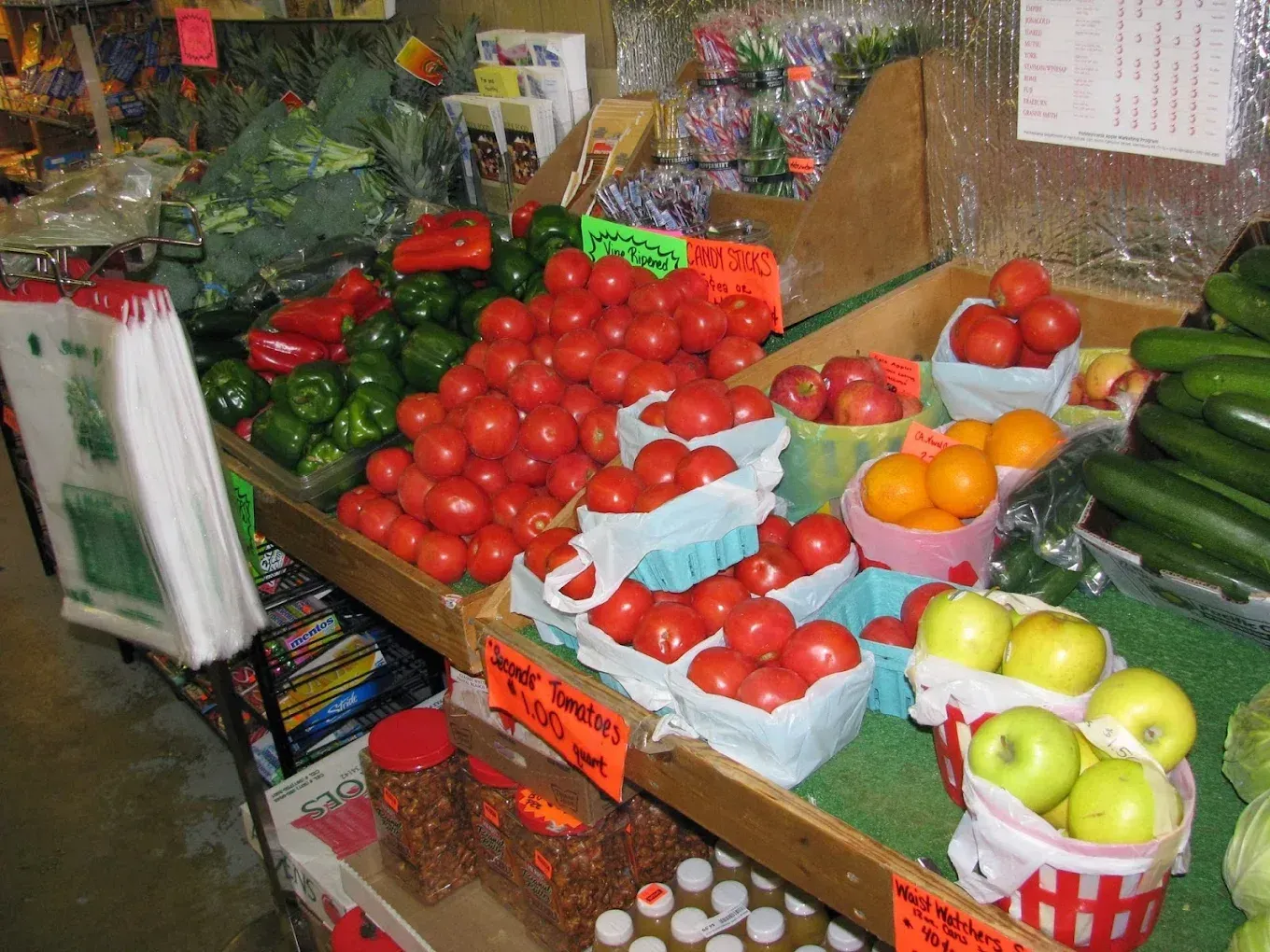 Fresh produce display with tomatoes, peppers, oranges, apples, and cucumbers at a grocery store market stall.