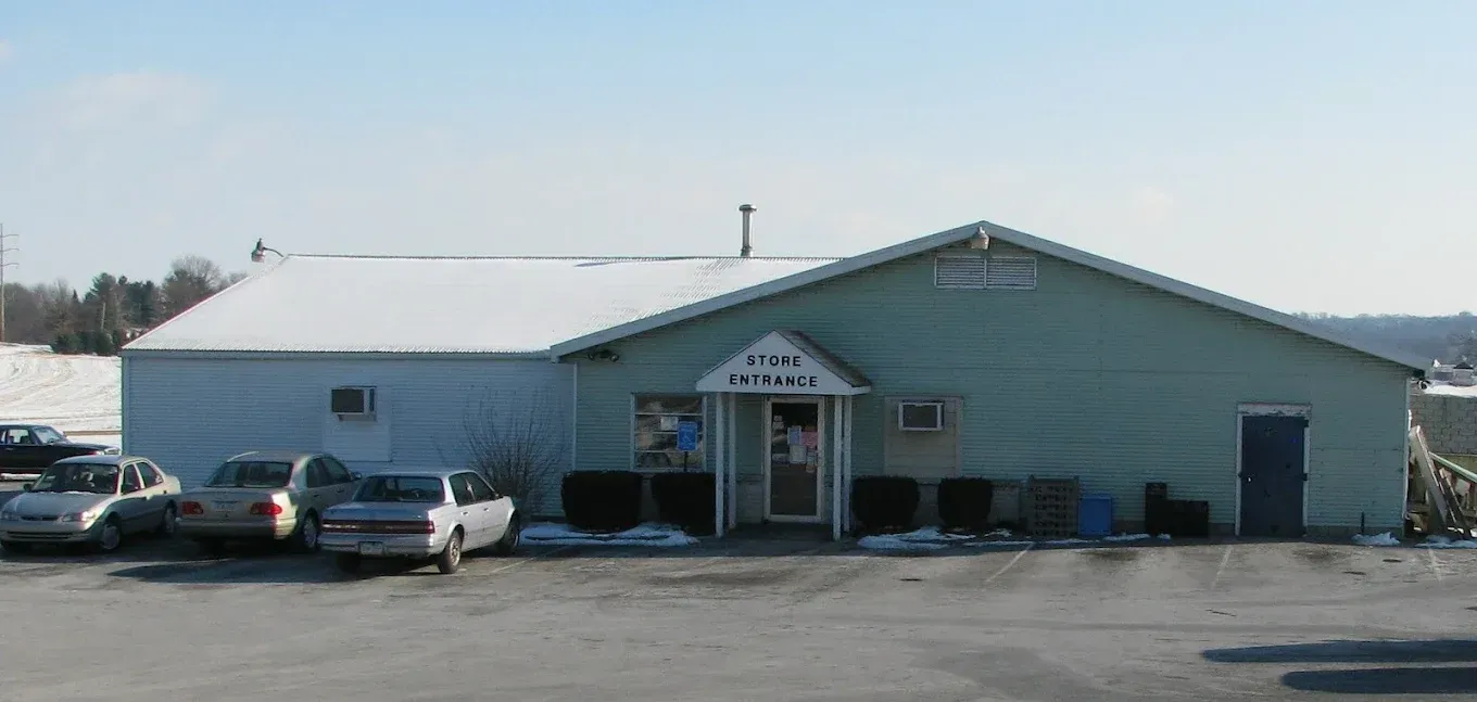 A light green, single-story building with a snow-covered roof and a central entrance, parked cars in a gravel lot.