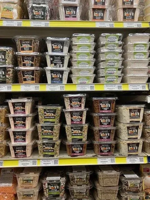 A grocery store shelf filled with various plastic containers of nuts, seeds, and grains arranged in neat rows and stacks.