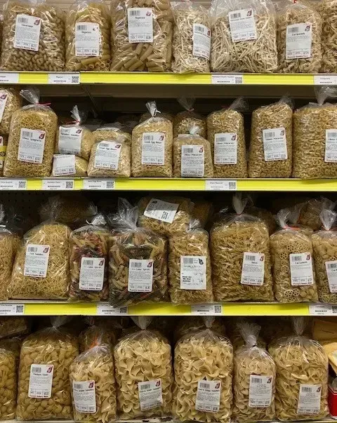 Shelves in a store stocked with rows of various shapes of pasta packaged in clear plastic bags.