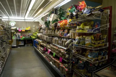 Shelves filled with various packaged snacks and goods line a narrow aisle in a grocery store under bright lighting.