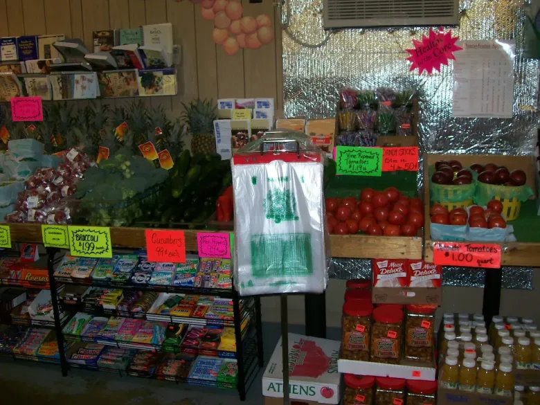 A produce market stand with shelves of fruits, vegetables, and snacks, featuring signs and plastic bags on a stand.