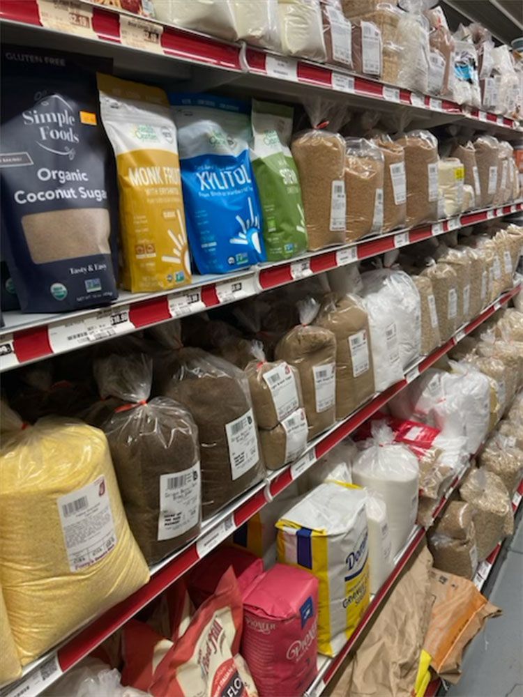 Shelves stocked with various bags of flour, sugar, and sweeteners in a grocery store aisle.