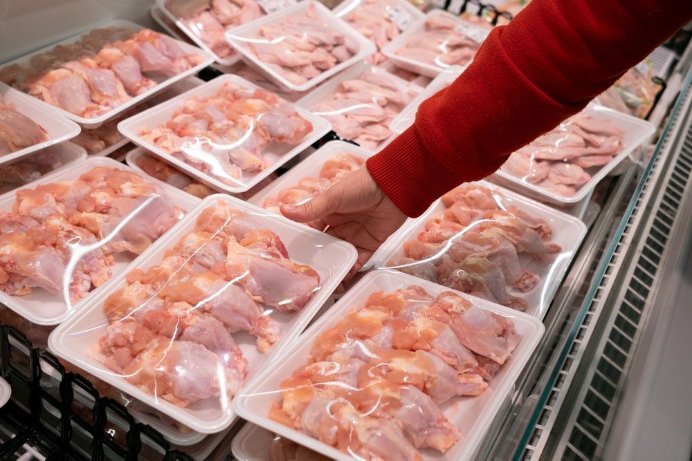 A hand in a red sleeve reaches for a plastic-wrapped package of raw chicken pieces in a grocery store refrigerator case.