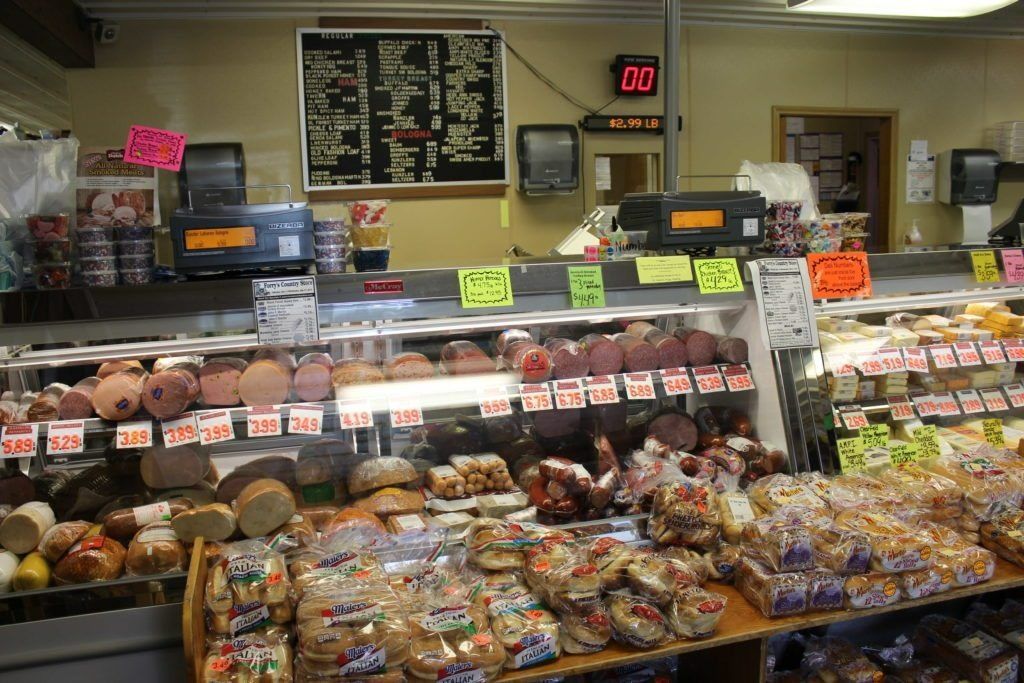 A well-stocked deli counter featuring various meats, cheeses, and baked goods behind glass displays under a menu board.