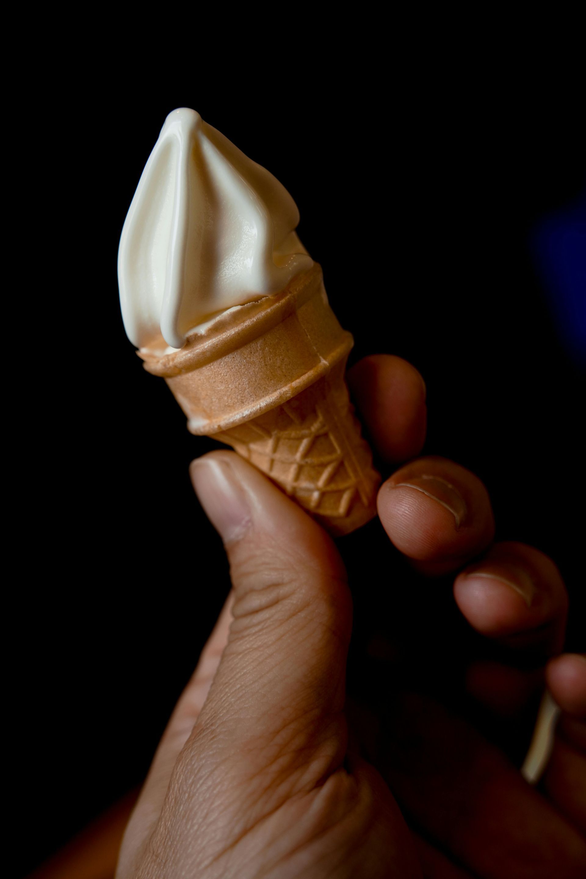 A hand holds a small soft-serve ice cream cone against a dark background.