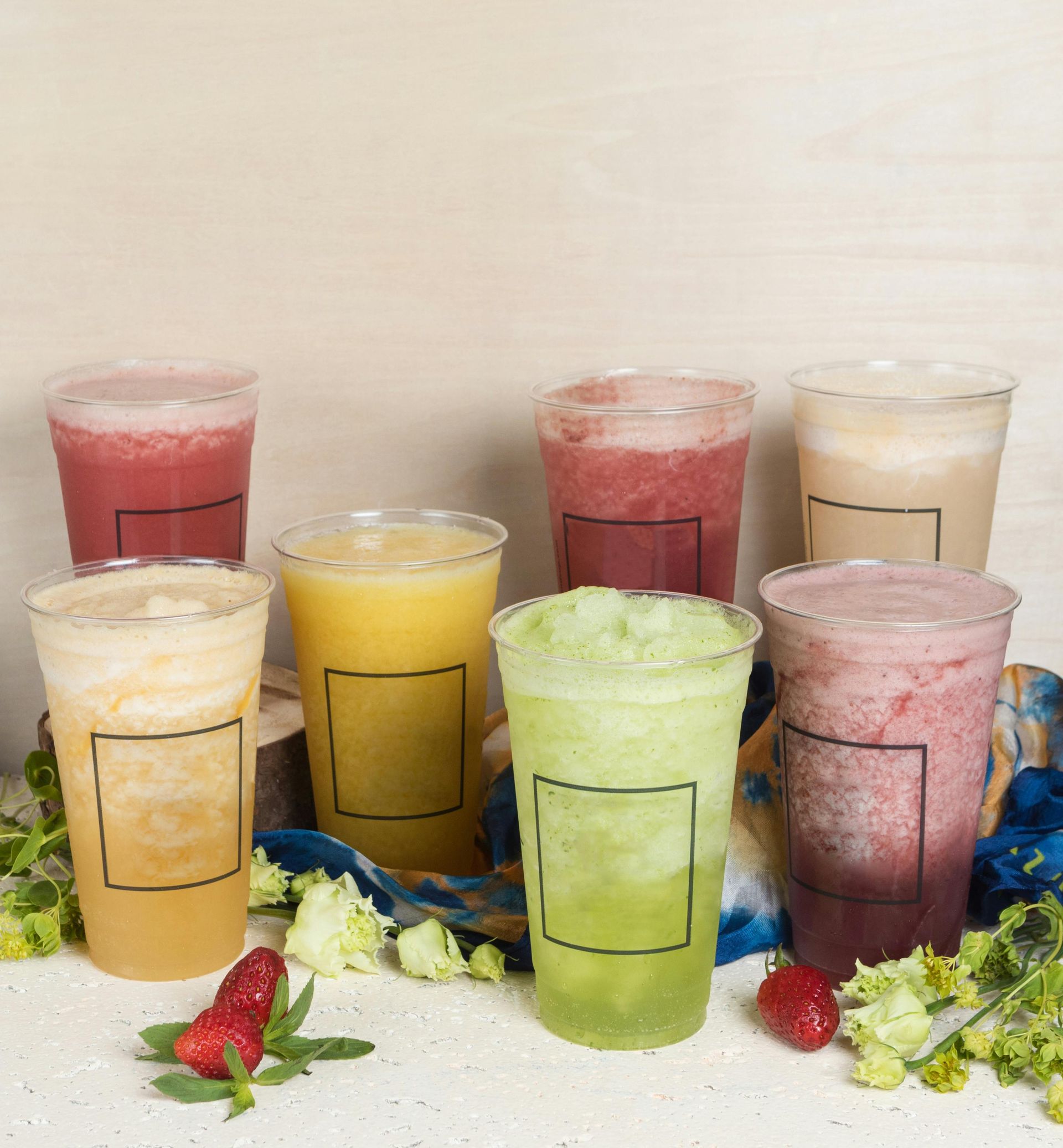 Seven clear plastic cups filled with colorful blended fruit drinks are arranged on a white table with fresh fruit.