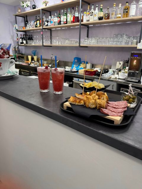 Bar con bevande e cibo sul bancone. Mensole con bottiglie e bicchieri. Due drink rossi e un vassoio di snack.