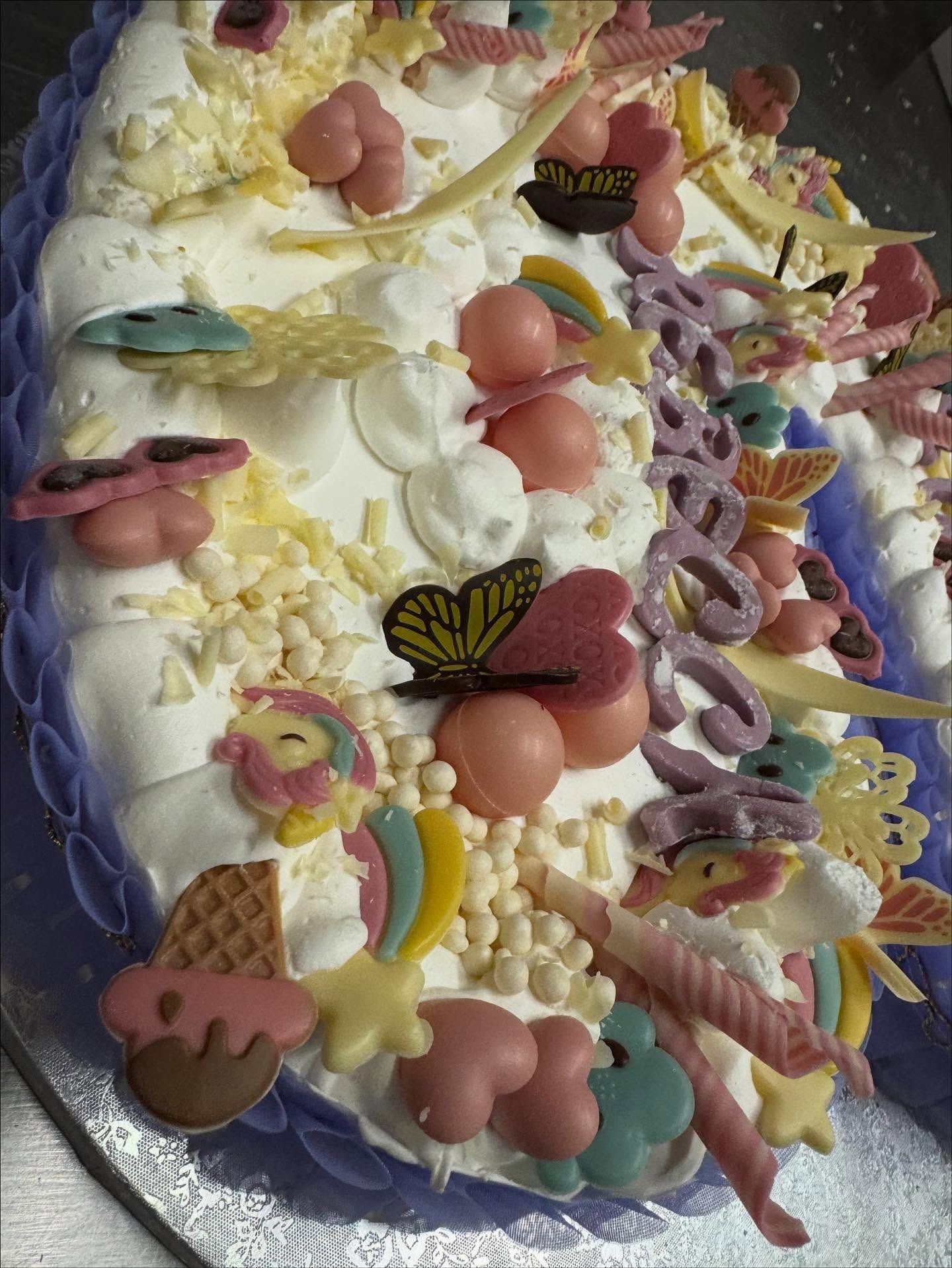 Torta decorata con caramelle color pastello, glassa e farfalle di cioccolato, su una superficie argentata.