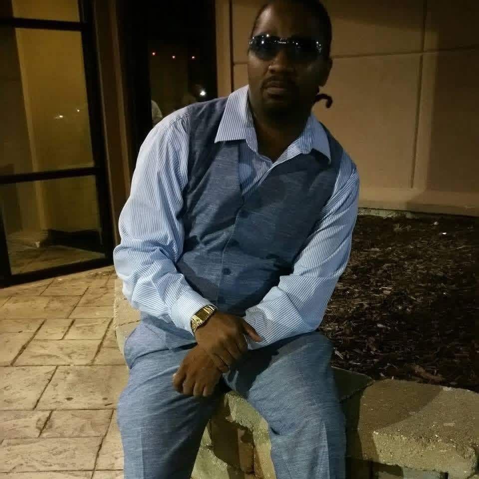 Man sitting on a stone ledge at night, wearing sunglasses, a light blue shirt, and denim overalls.