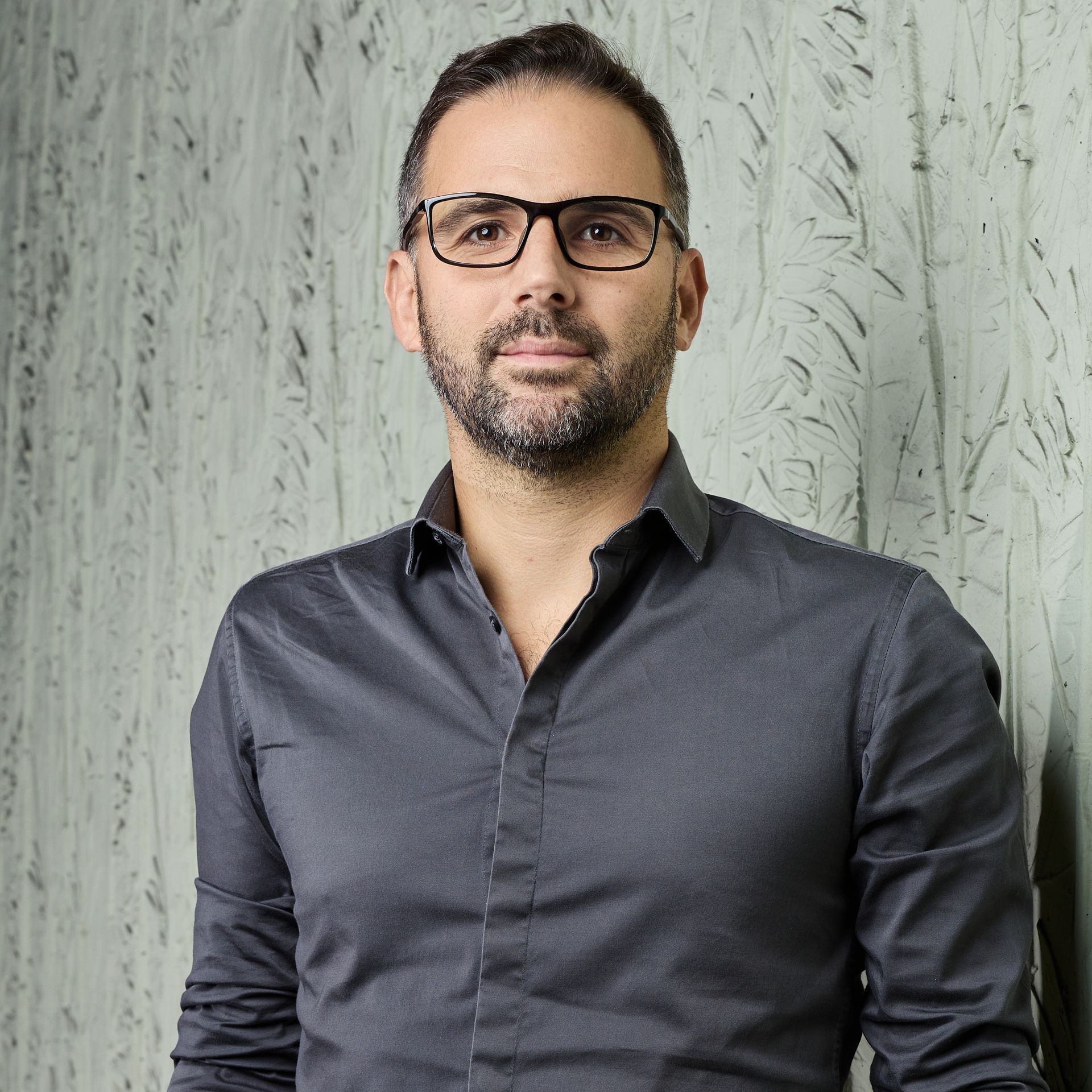 Un homme en chemise gris foncé et lunettes se tient debout contre un mur texturé.