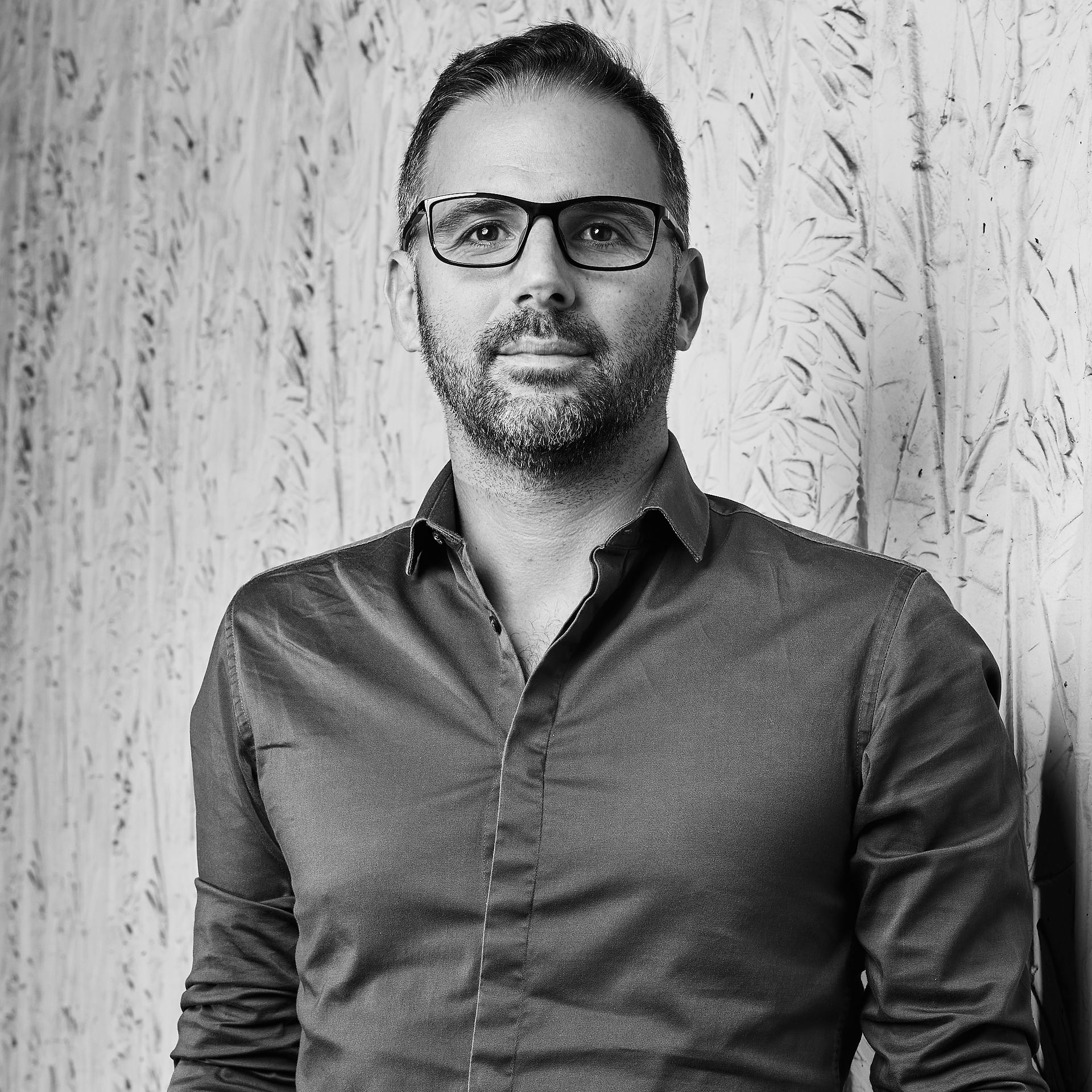 Portrait d'un homme portant des lunettes et une chemise sombre, debout devant un mur texturé.