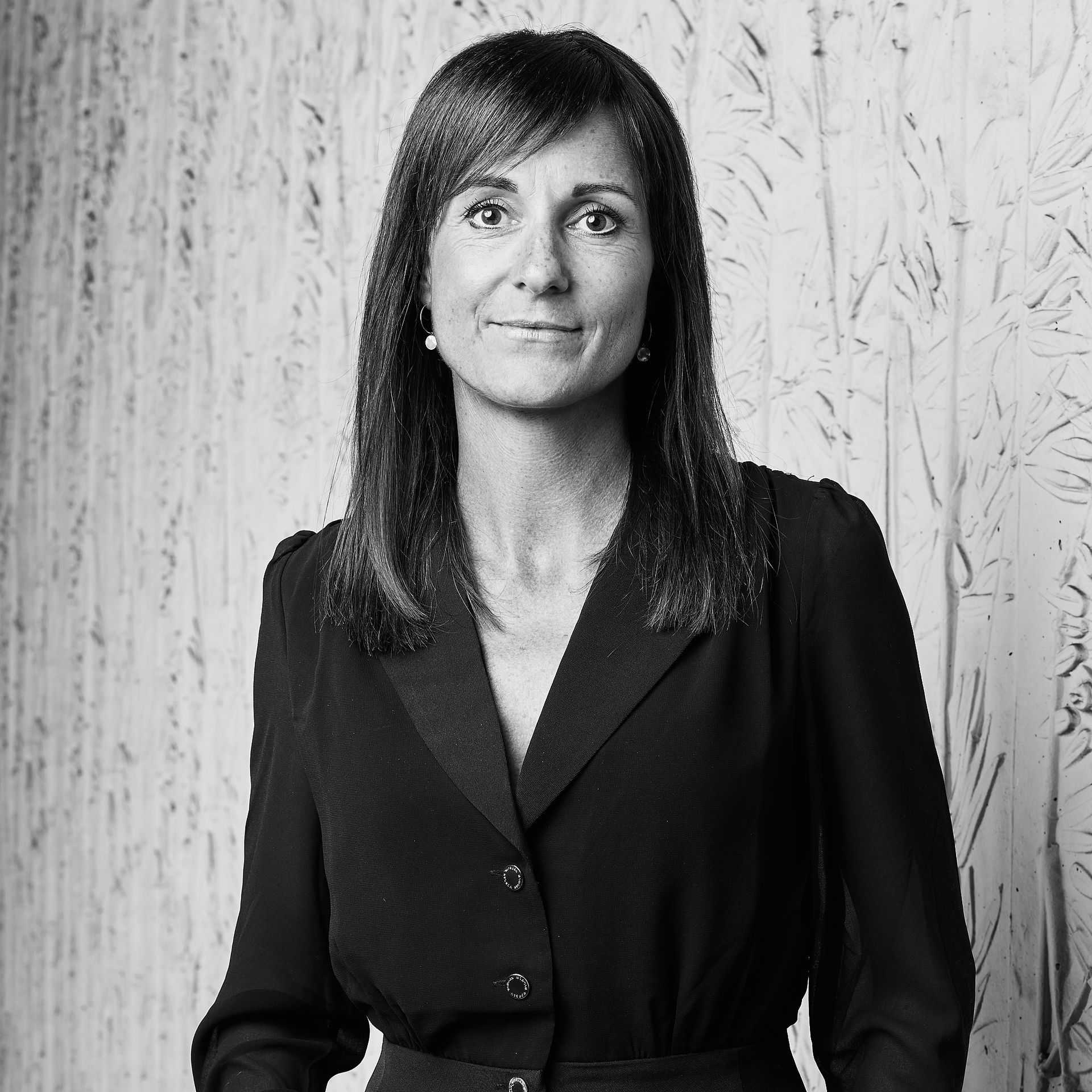 Portrait d'une femme en blazer foncé, debout devant un mur lambrissé clair.