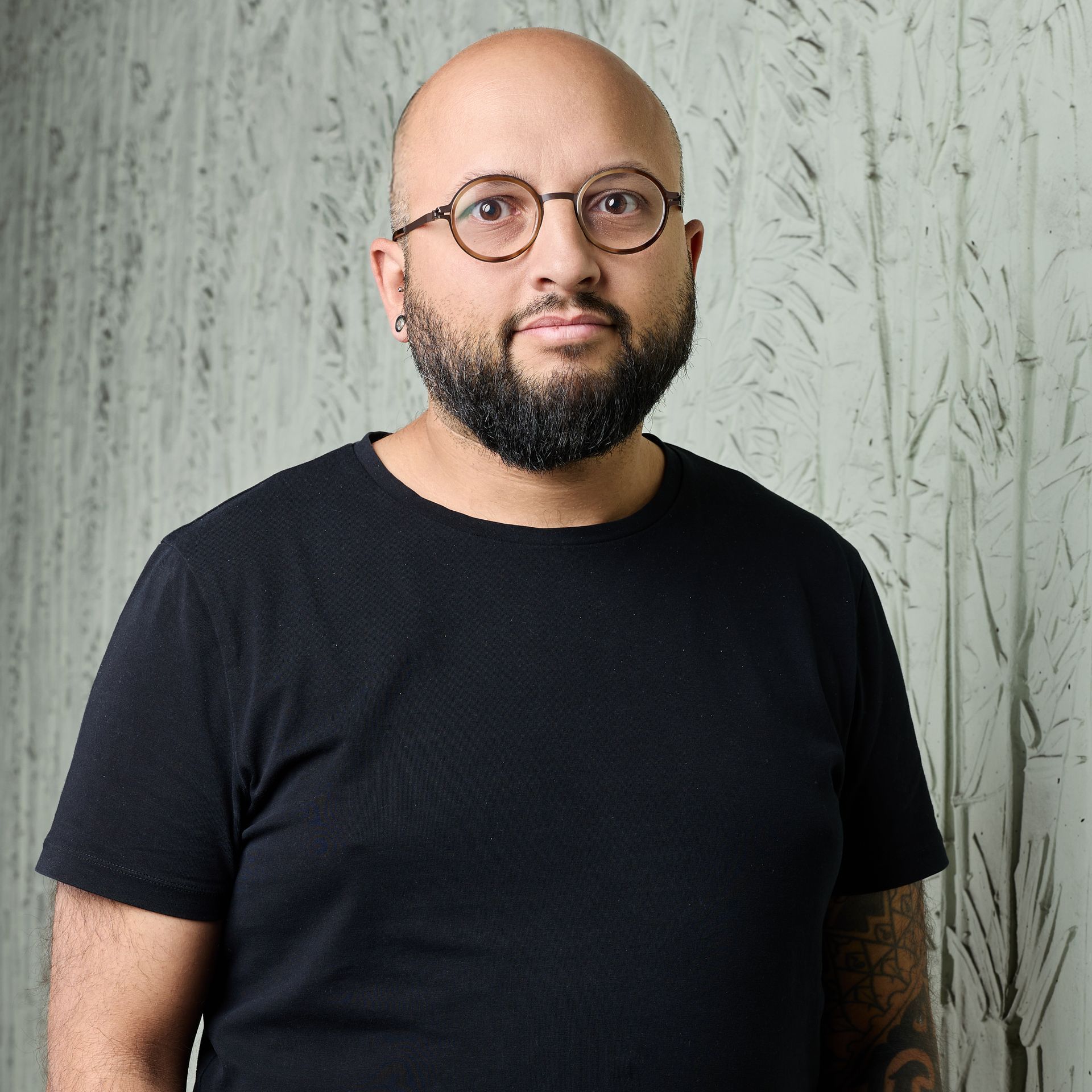 Homme portant des lunettes et une barbe, vêtu d'un t-shirt noir, debout contre un mur gris texturé.